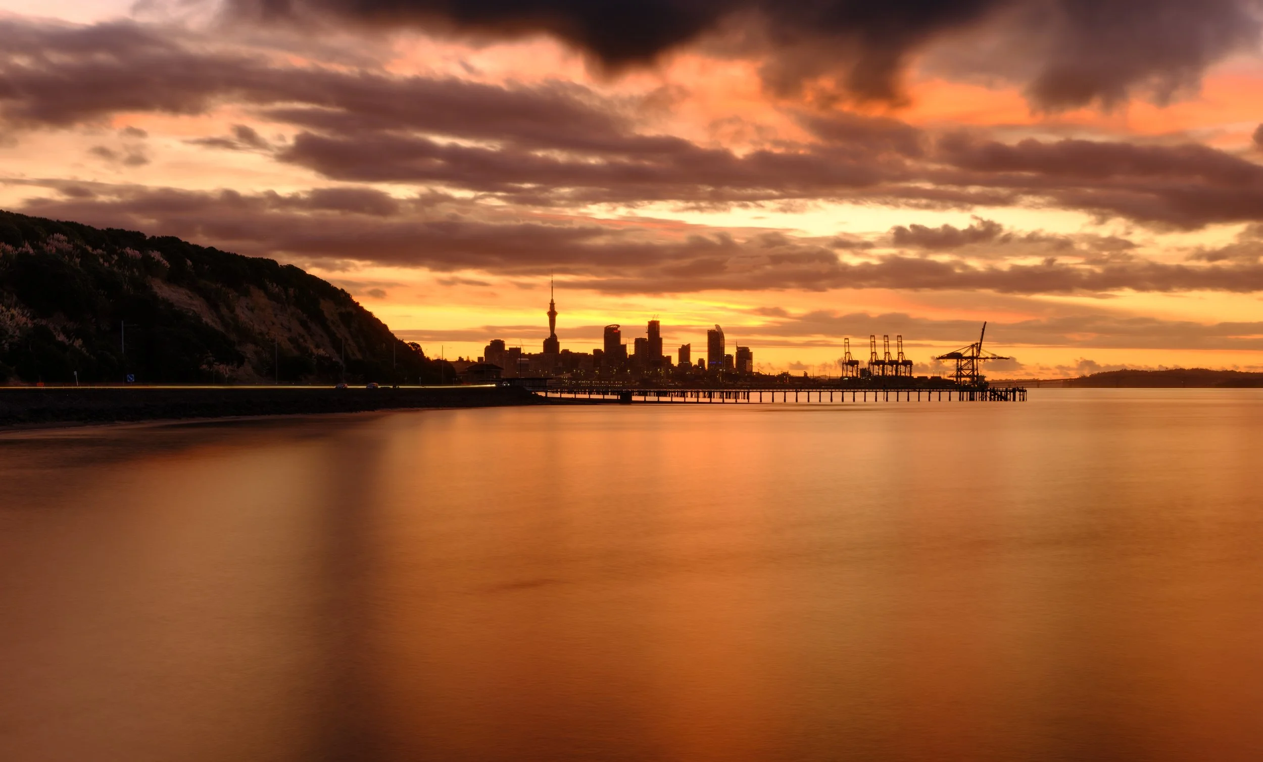 Sunset over a city skyline with a pier and industrial cranes, reflected in calm water, with dark clouds overhead.