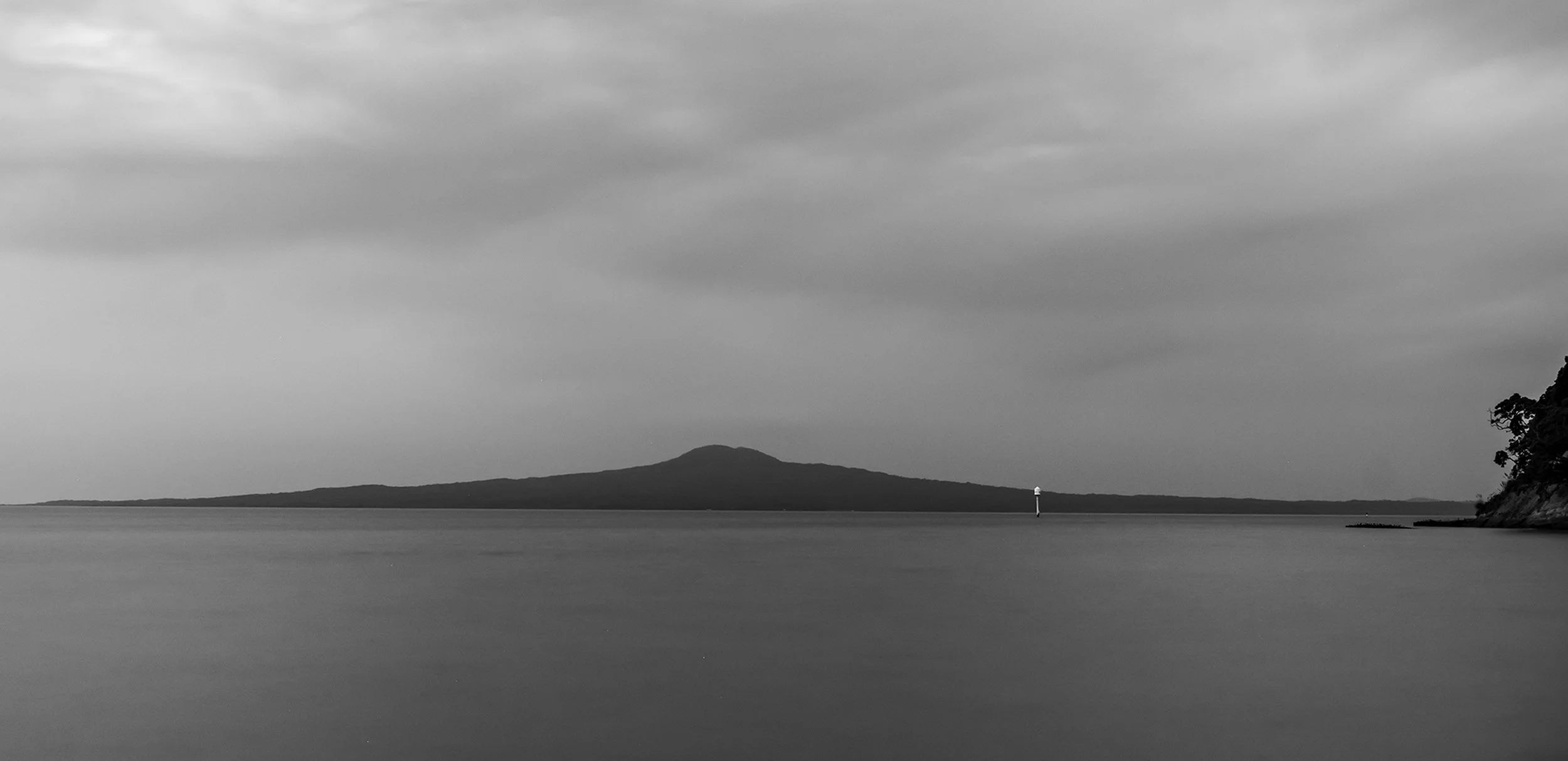 A black-and-white photo of a calm body of water with a distant mountain or island in the background, a lighthouse near the horizon, and cloudy sky above.