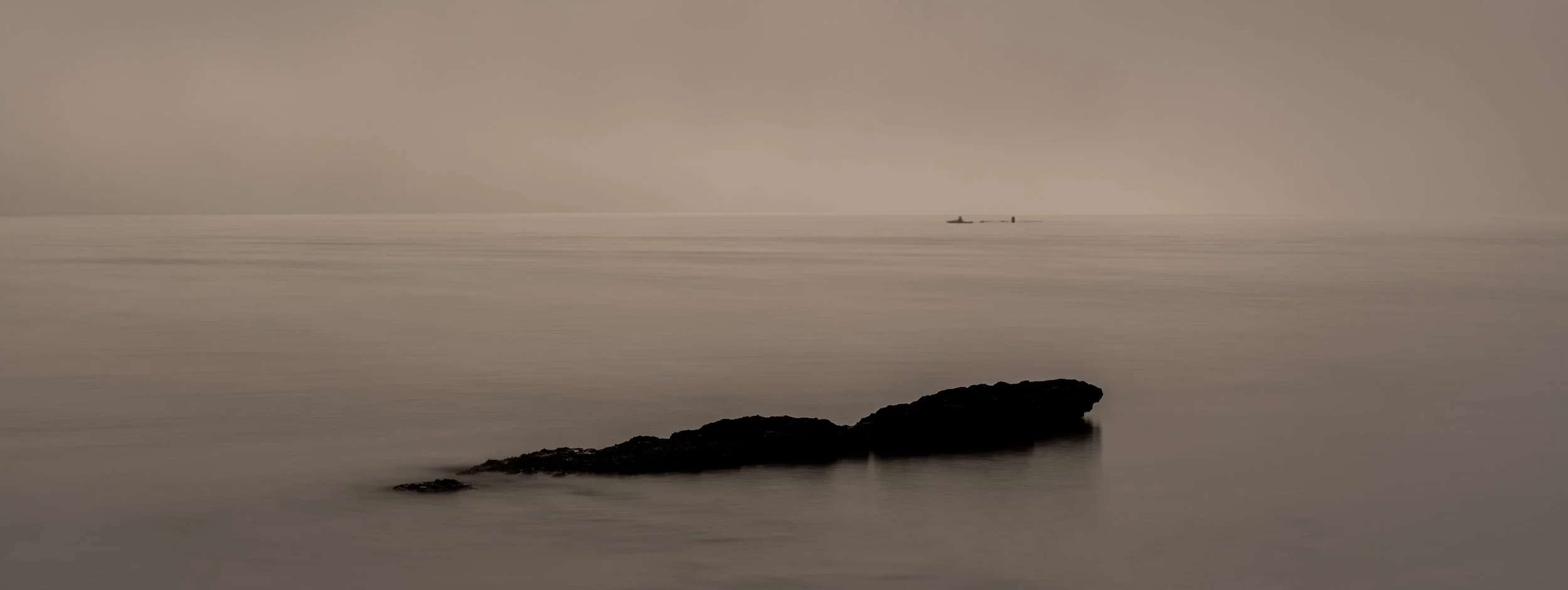 Seascape with a large rock in the foreground, calm water, and a small boat in the distance under an overcast sky.