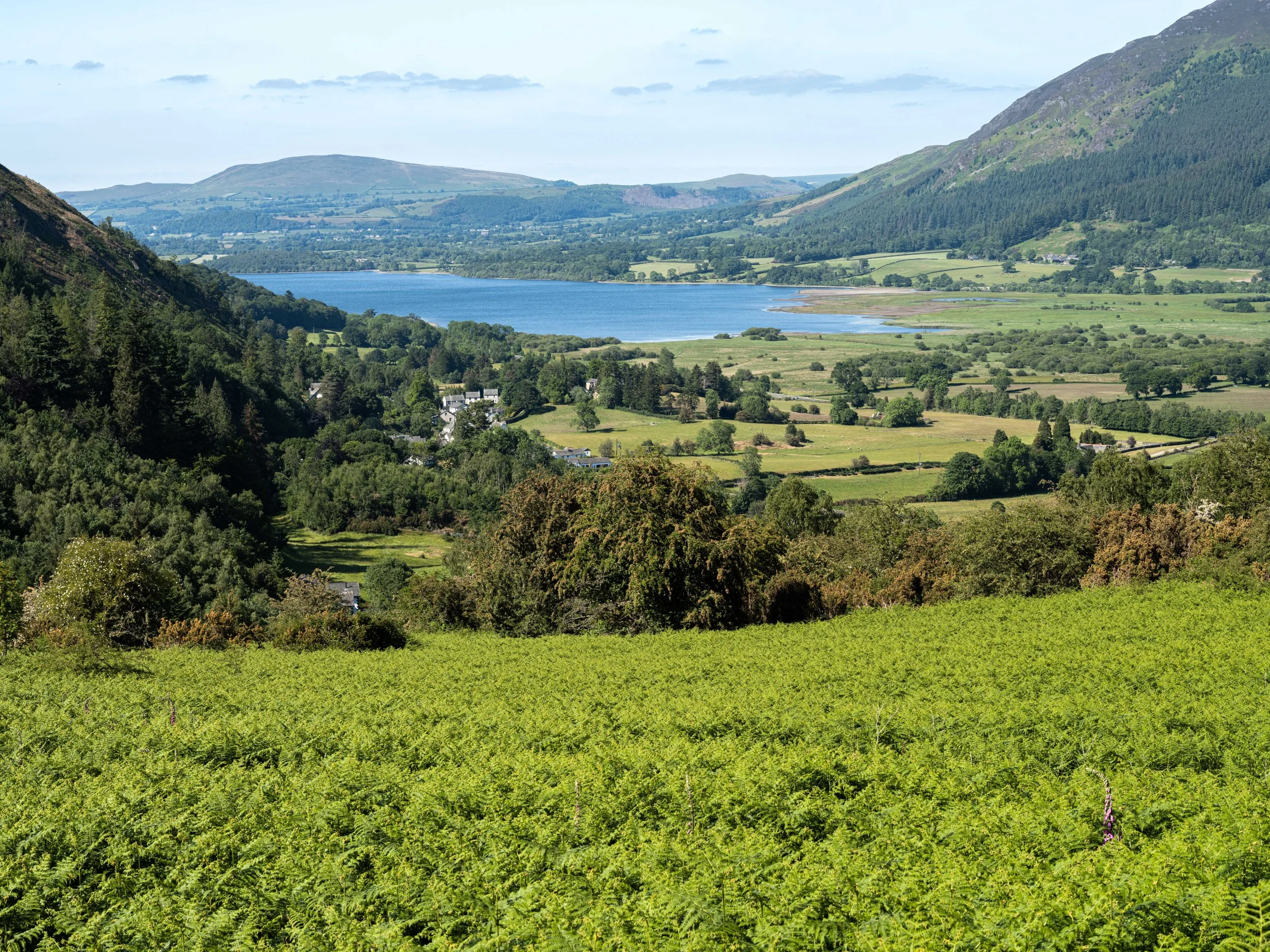 Lush green valleys with a lake in the distance surrounded by mountains under a partly cloudy sky.