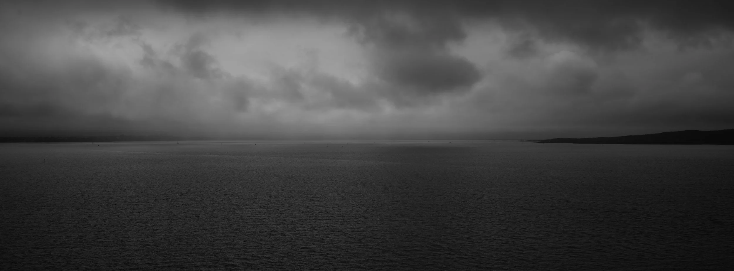 Black and white photo of a large body of water under a cloudy sky, with distant land on the horizon.
