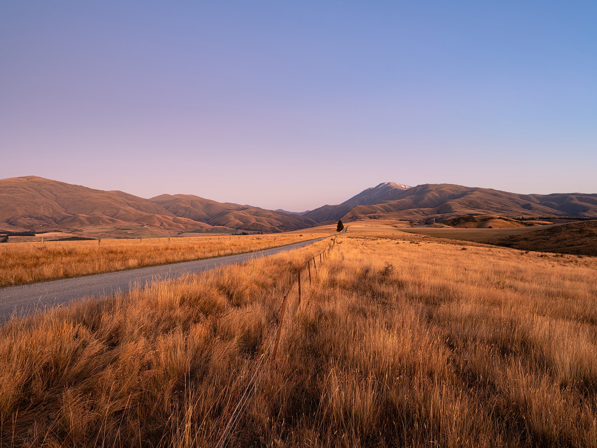 Open countryside with rolling hills, a dirt road, and tall golden grass under a clear blue sky.