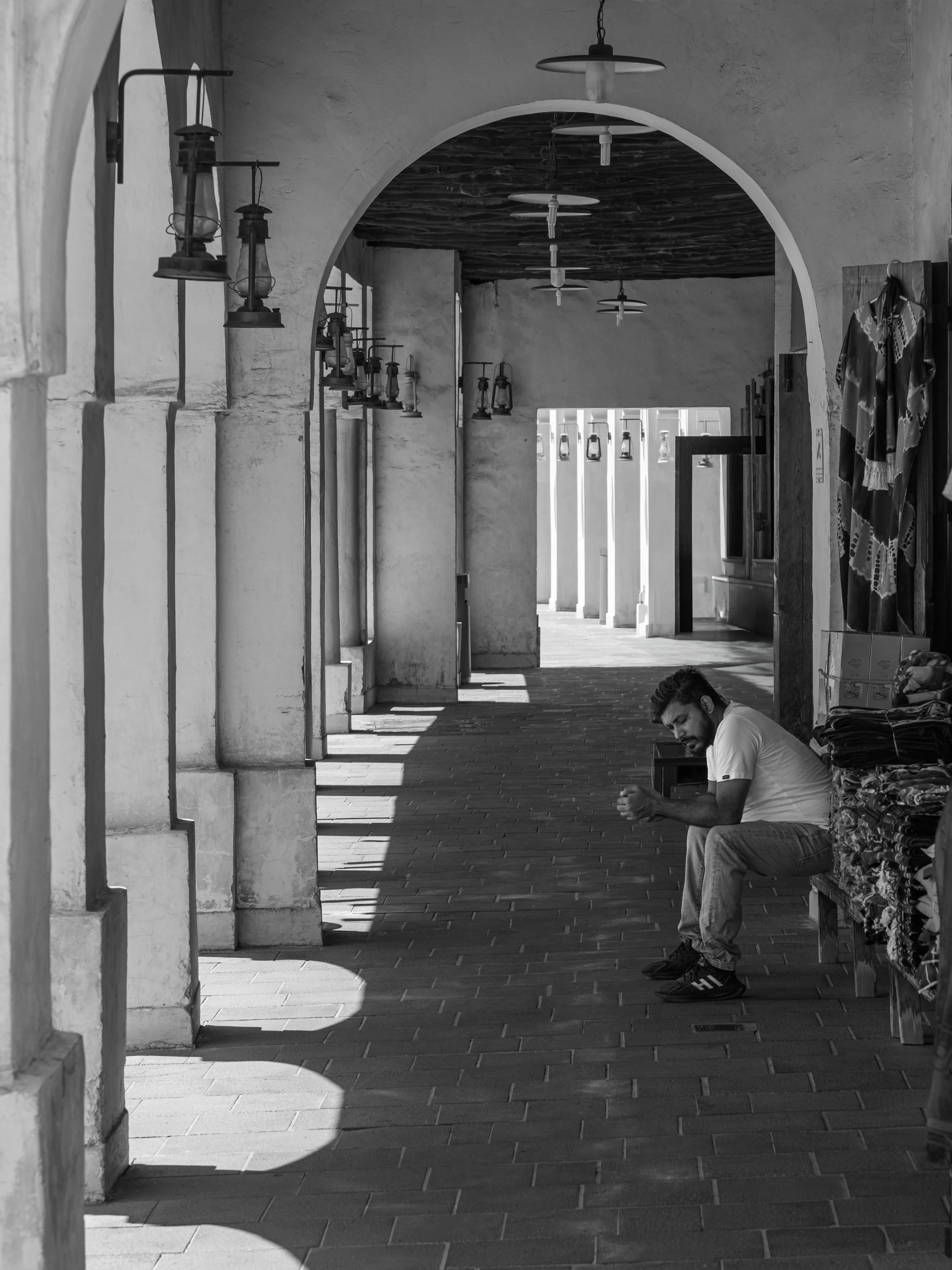 A man sitting on a bench under an arched walkway with vintage lanterns attached to the wall, shadows cast on the ground, and a cloth display on the right.