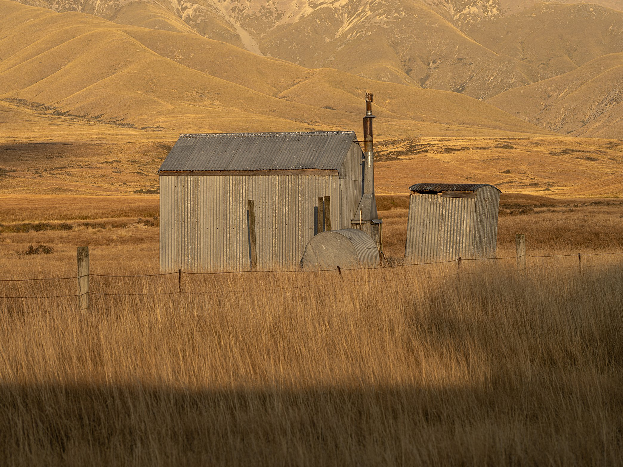 An old, corrugated metal building in a grassy field with rolling hills in the background during daytime.