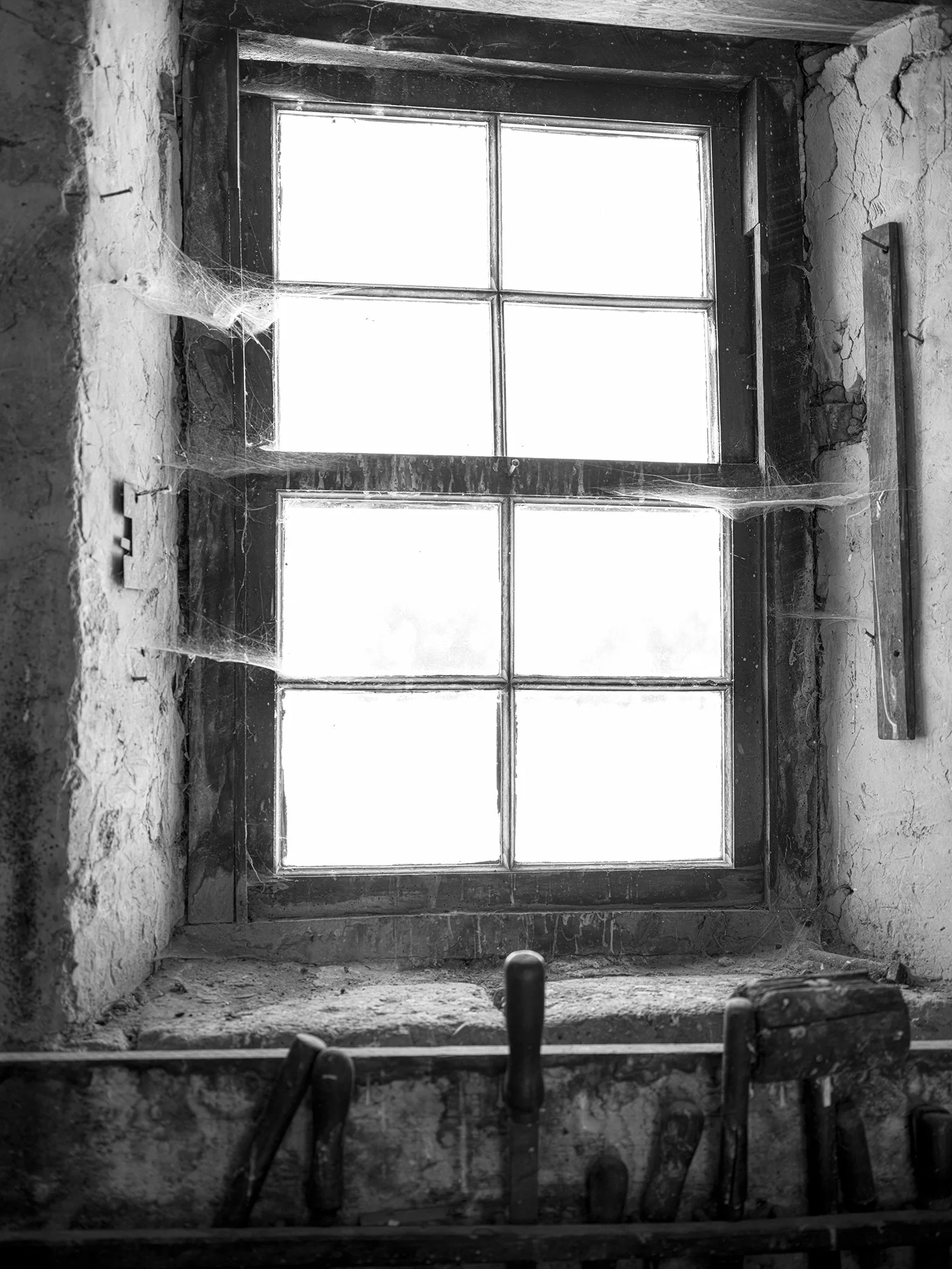 Black and white photo of an old, rustic window with divided panes. Cobwebs are present in the corners, and an old tool is resting on the windowsill.