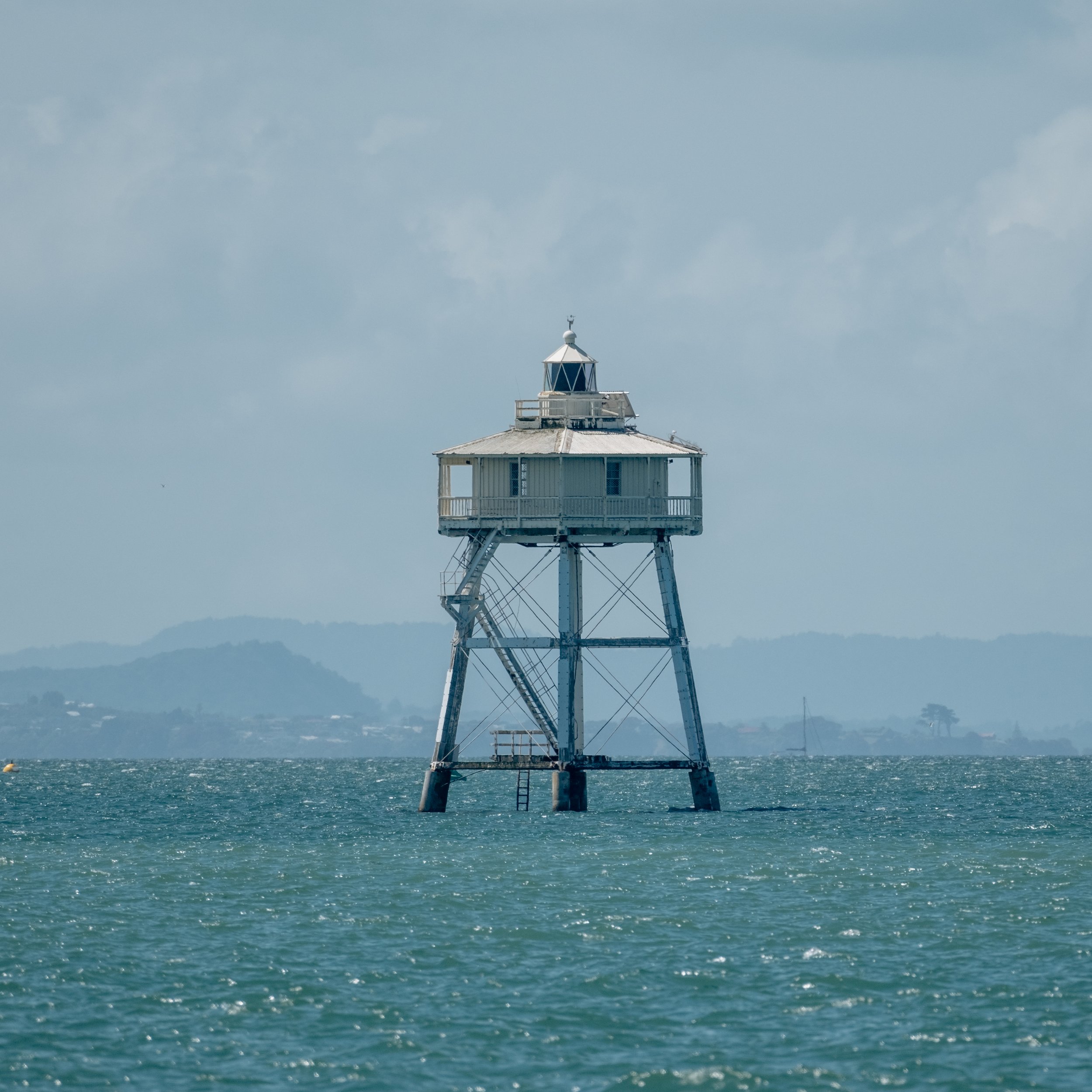 A tall lighthouse standing in the ocean with a mountainous coastline in the background and cloudy skies overhead.
