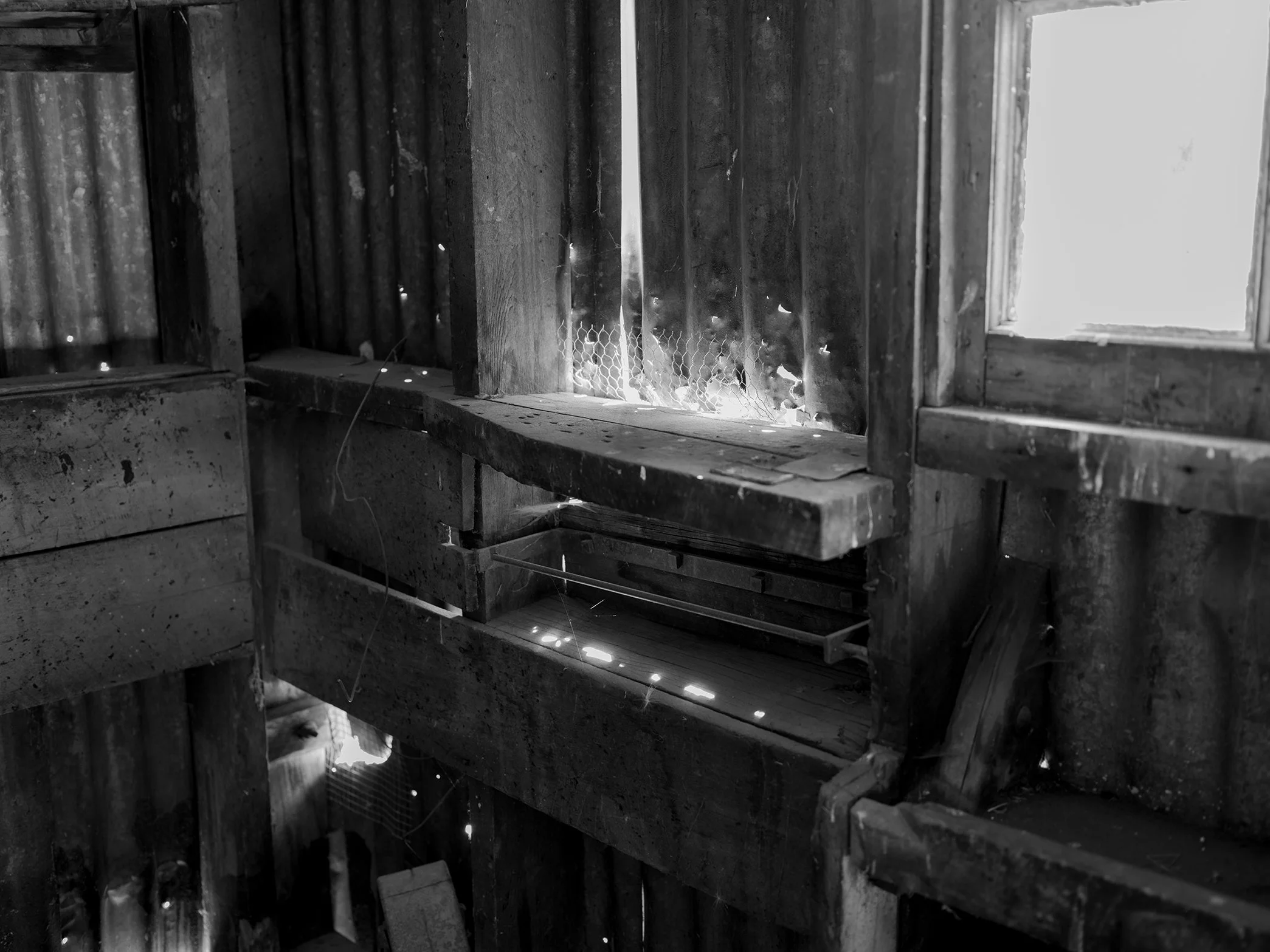 Interior of a weathered wooden structure with gaps letting in sunlight, showing a rustic, aged window.