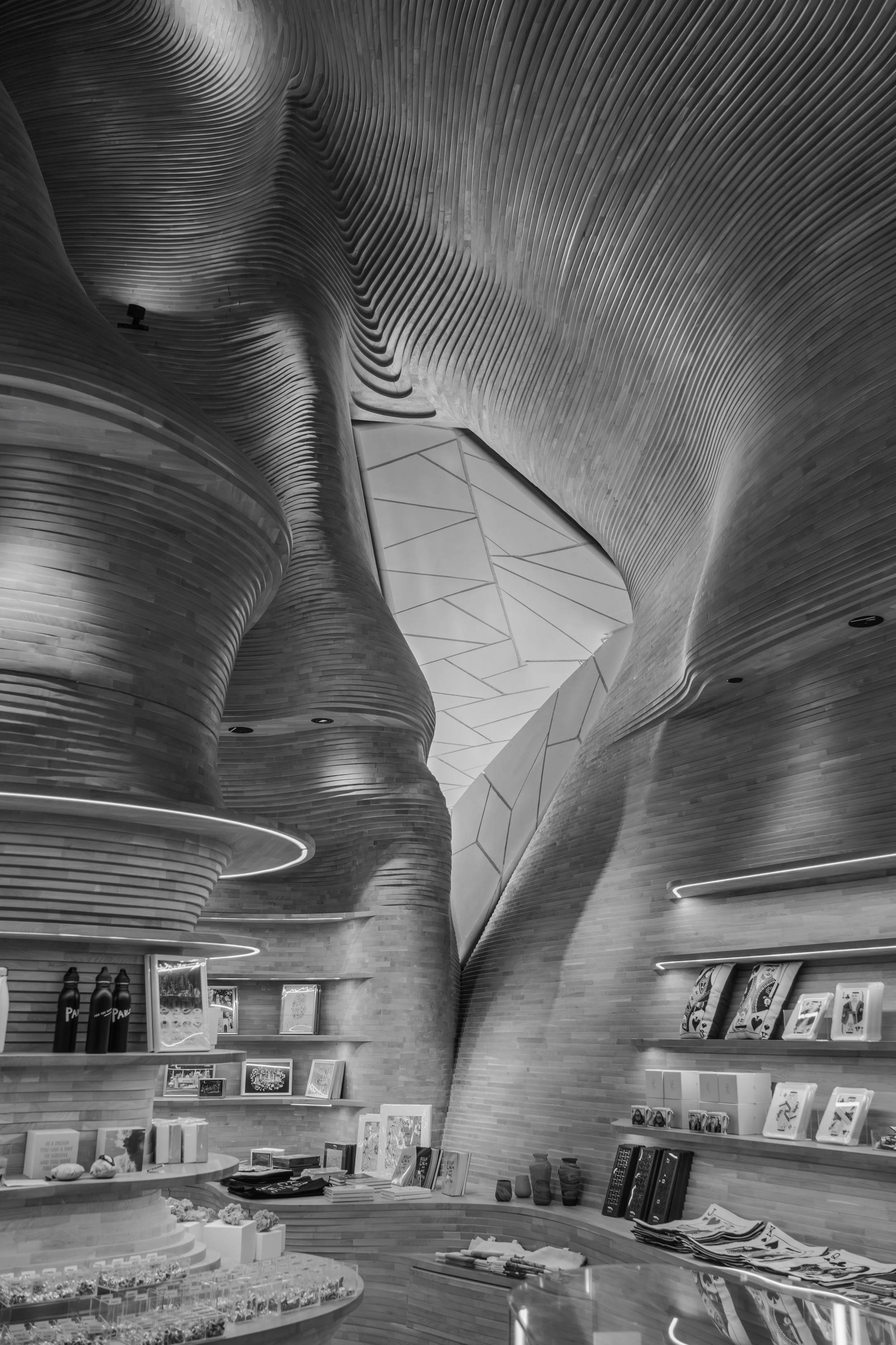 Interior of a modern store with curved wooden walls, displaying books, framed pictures, and bottles on shelves, with a geometric ceiling design.