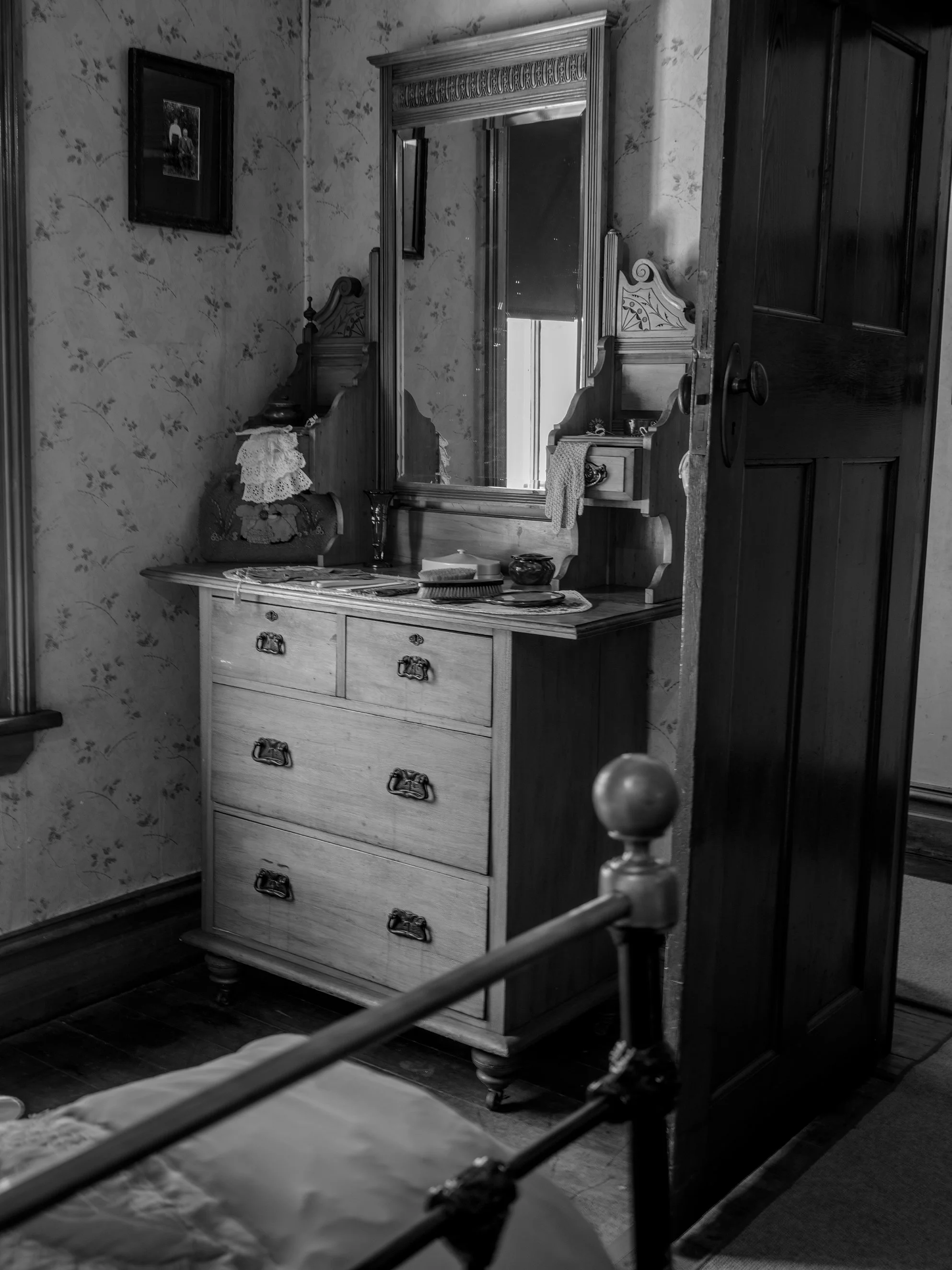 A black and white photo of a vintage bedroom interior featuring a wooden dresser with a mirror, a bed with metal posts, a floral wallpaper, and framed pictures on the wall.