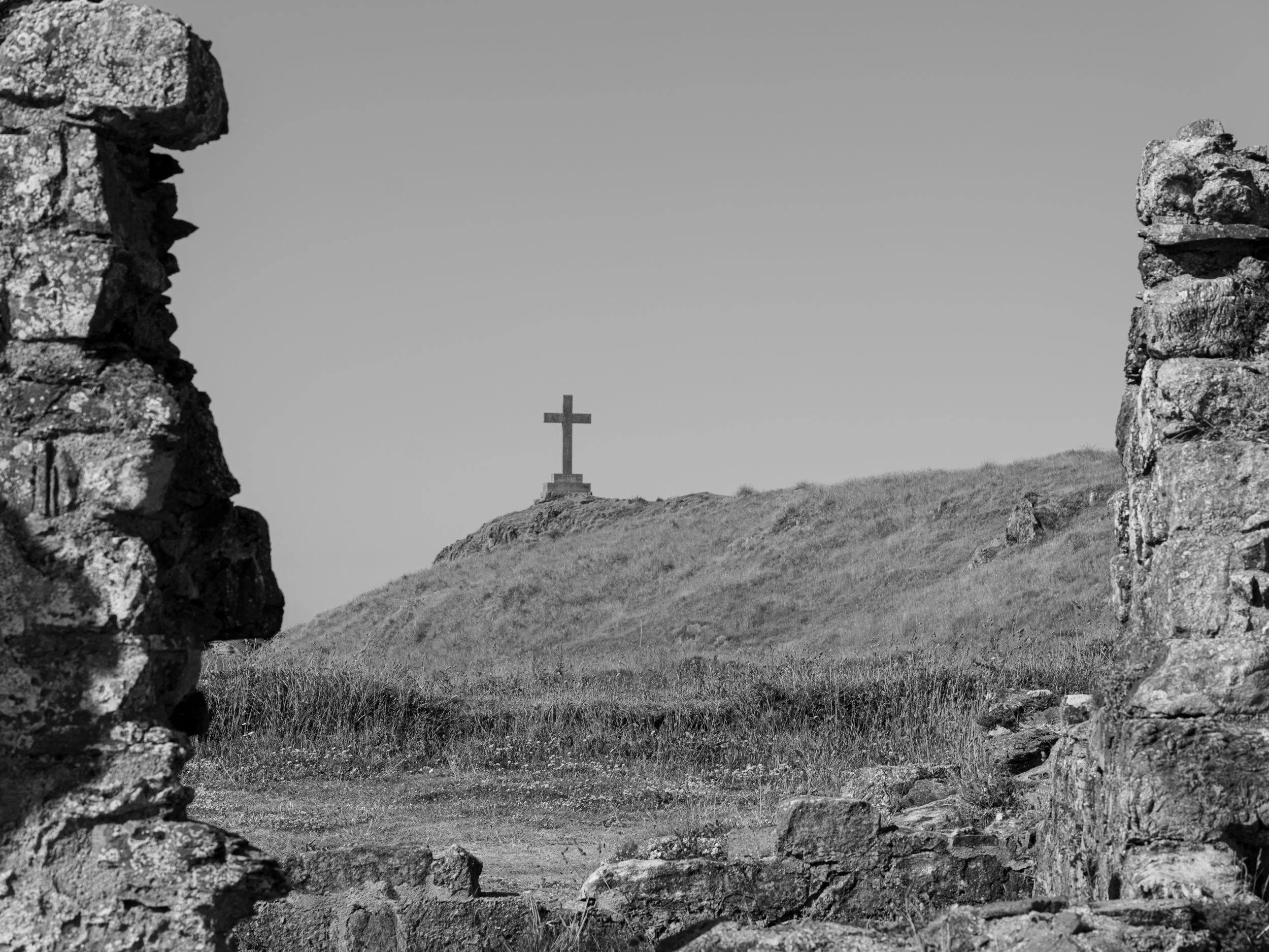 View through an opening in an old stone wall shows a hill with a large cross on top in the distance, under clear sky.