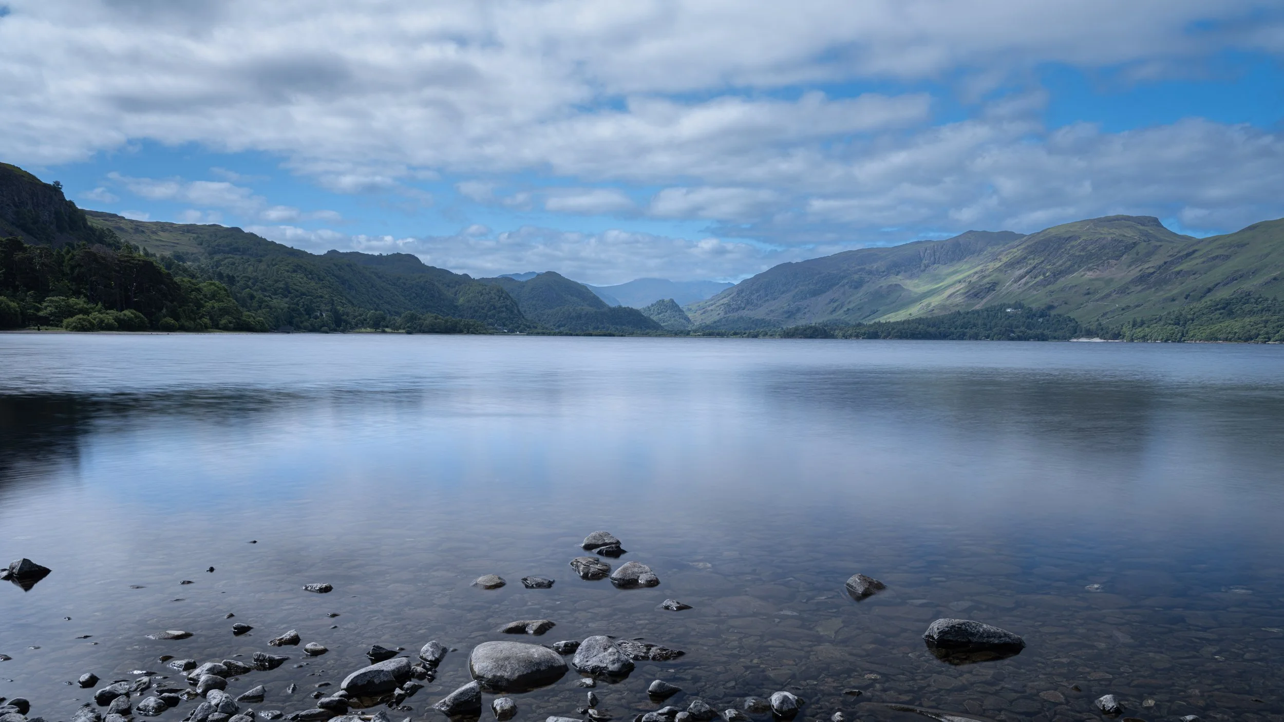 A calm lake surrounded by lush green mountains under a partly cloudy sky.