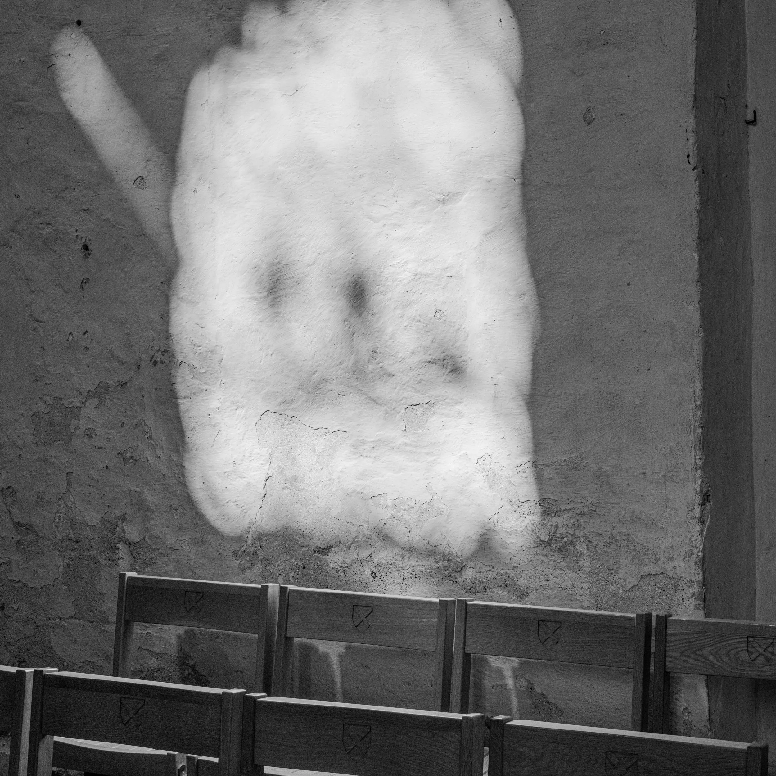 Black and white photograph of a large face mural on a textured wall, with a row of empty wooden chairs in the foreground.