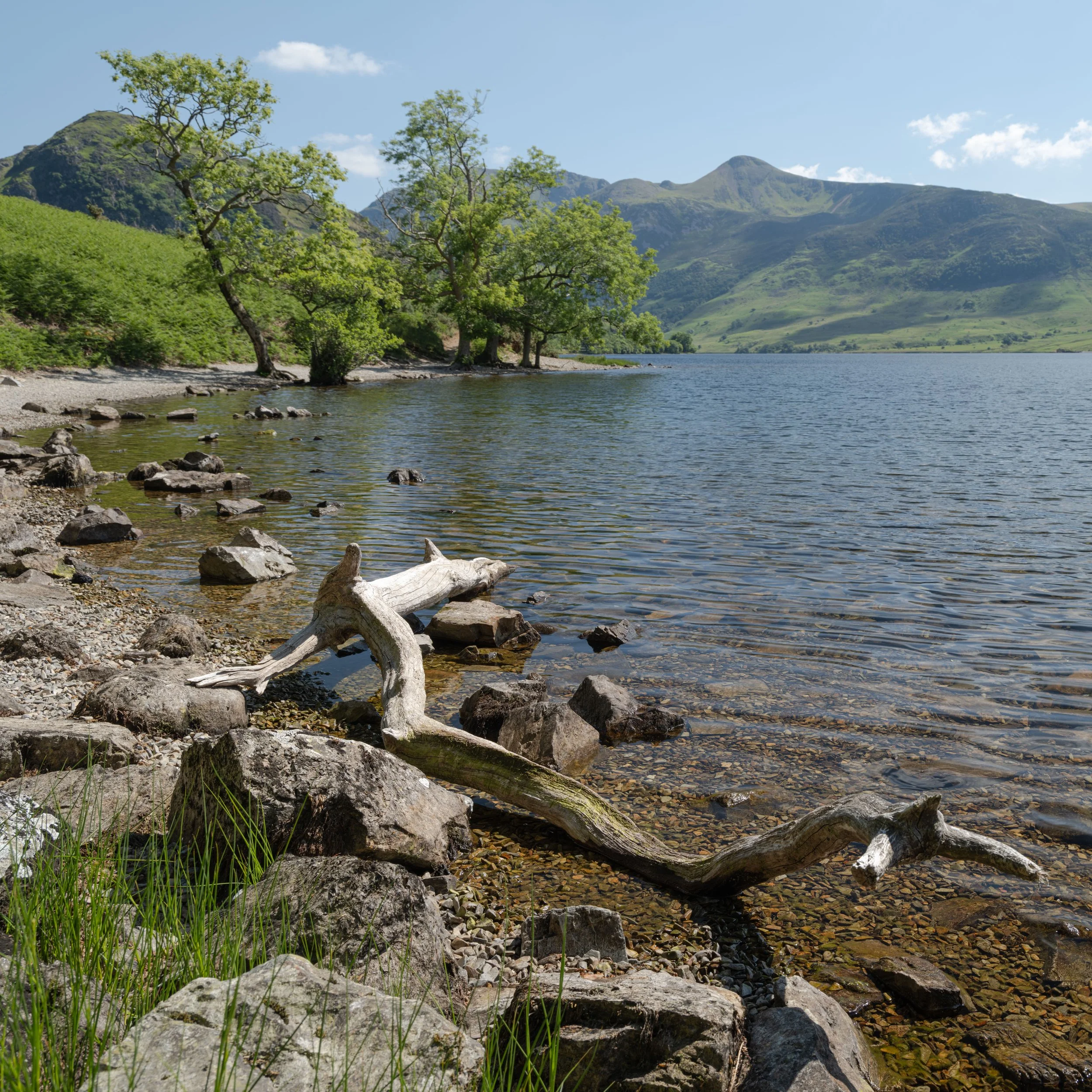Scenic view of a lake with clear water, surrounded by green trees and mountains in the background under a partly cloudy sky.