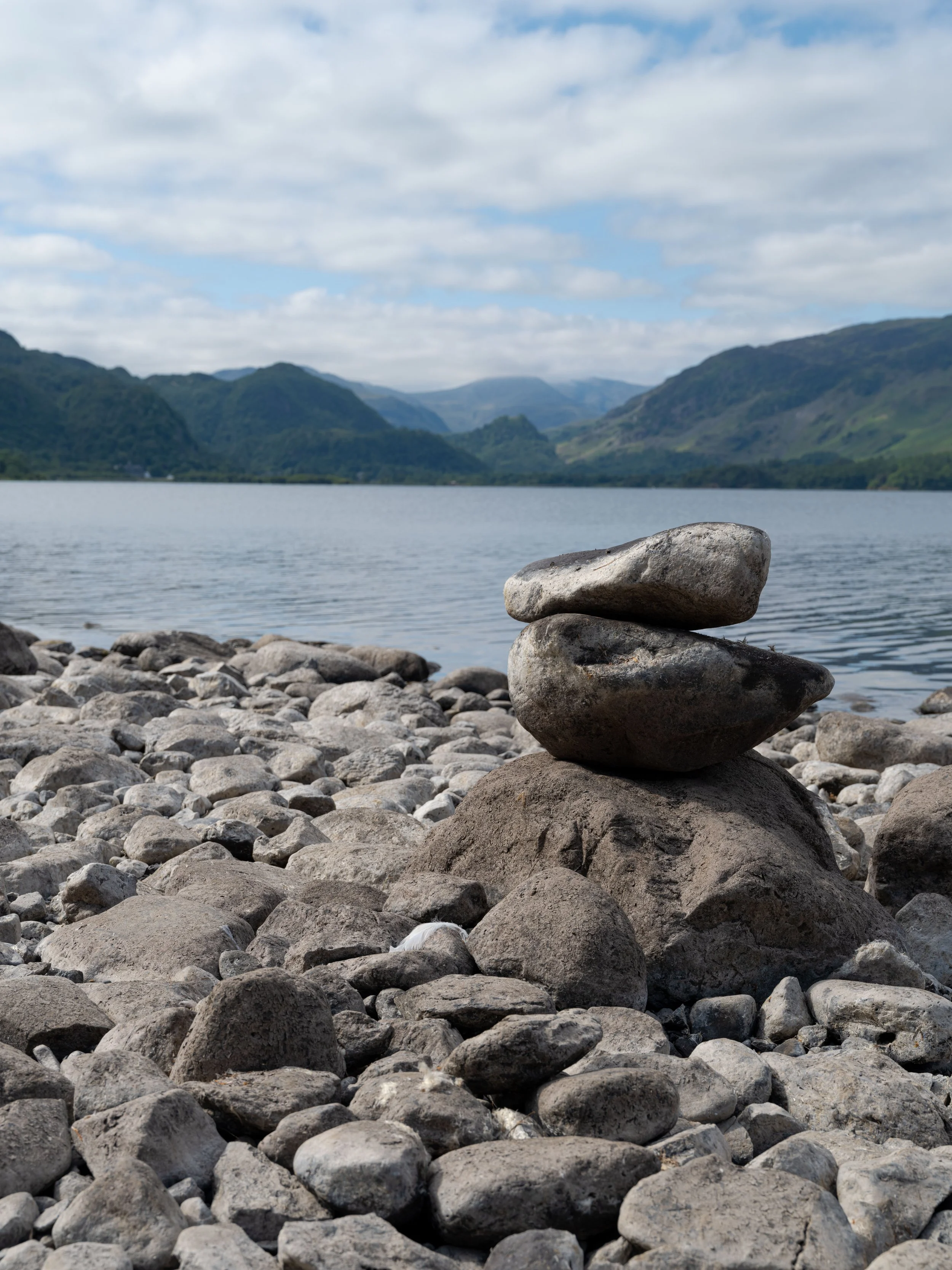 Stacked rocks on a rocky lakeshore with mountains and cloudy sky in the background.