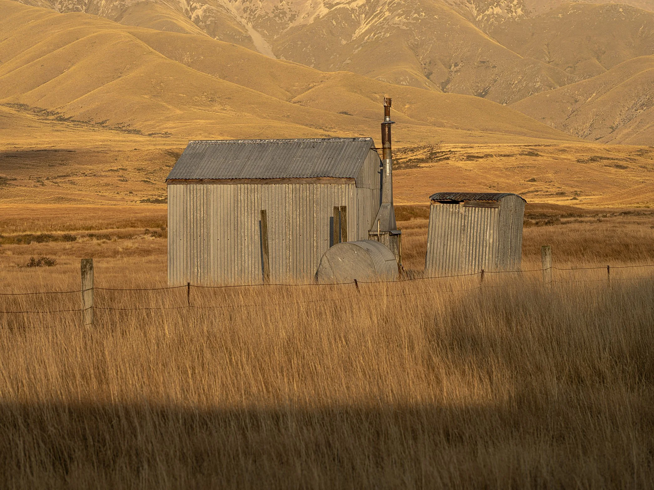 An old metal shed and silo in a grassy field with rolling hills and mountains in the background at sunset.