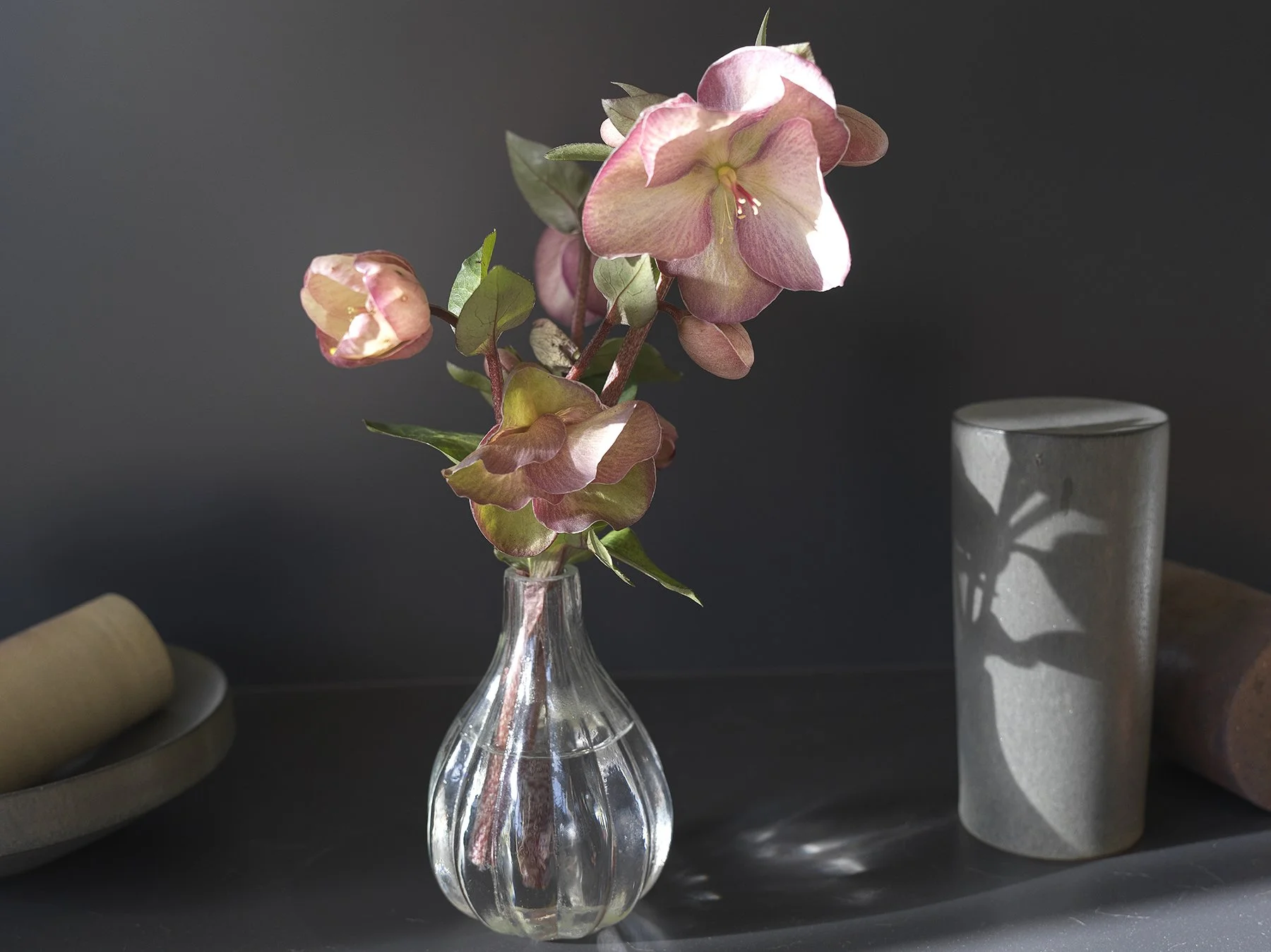 Pink and green flowers in a clear glass vase, with a gray cylindrical object and other objects on a dark surface.