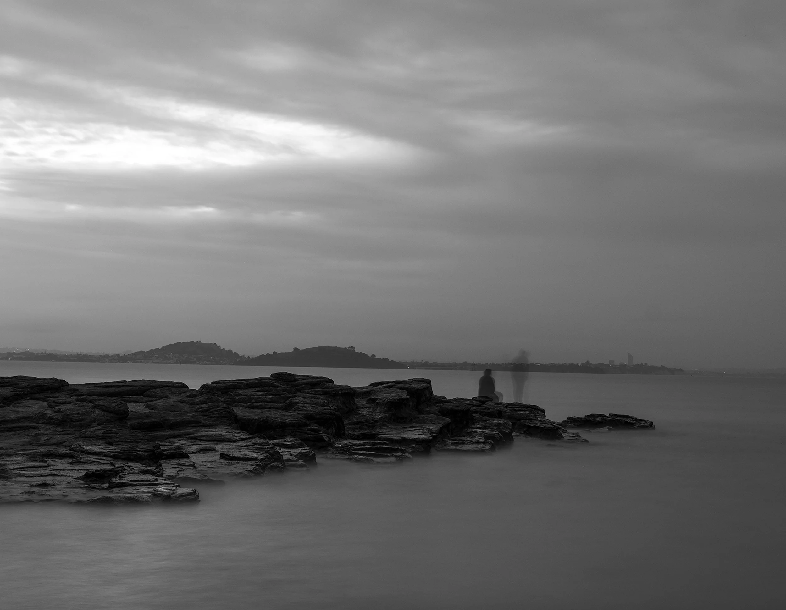Black and white photo of a person sitting on rocks by the water, with distant land and cloudy sky in the background.