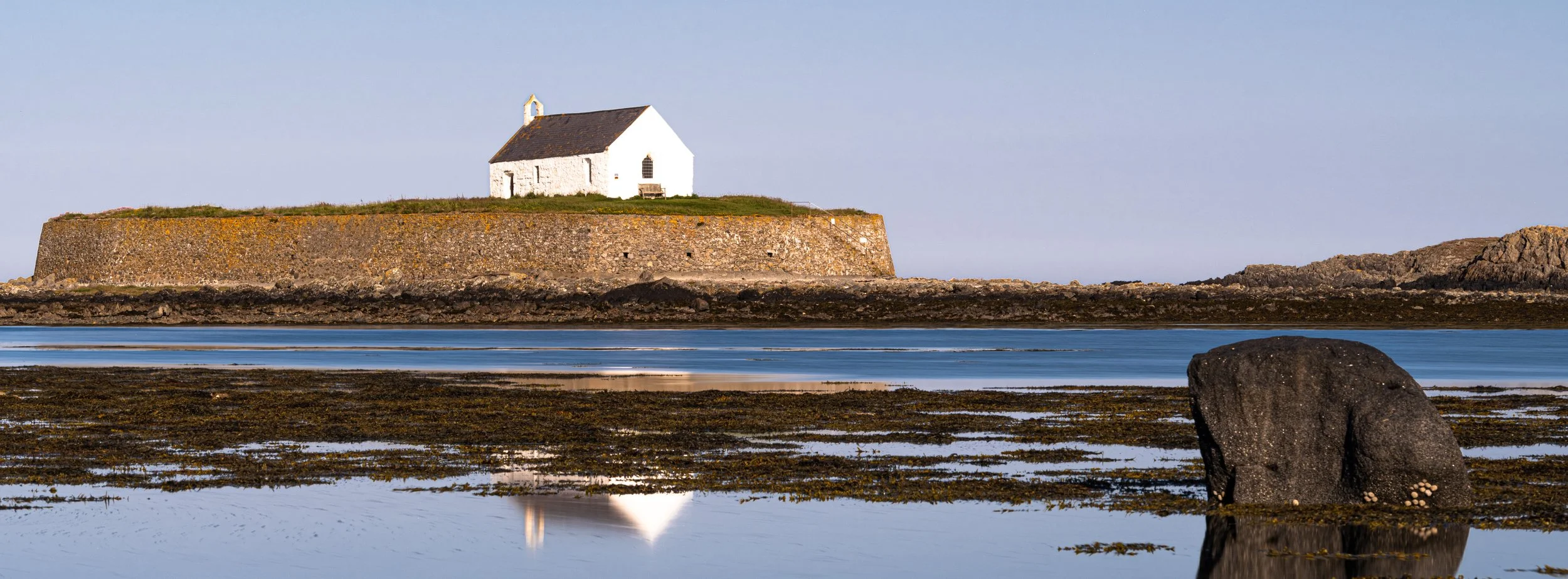 A small white church sitting atop a stone pier by the water, with rocks and tide pools in the foreground, under a clear sky.