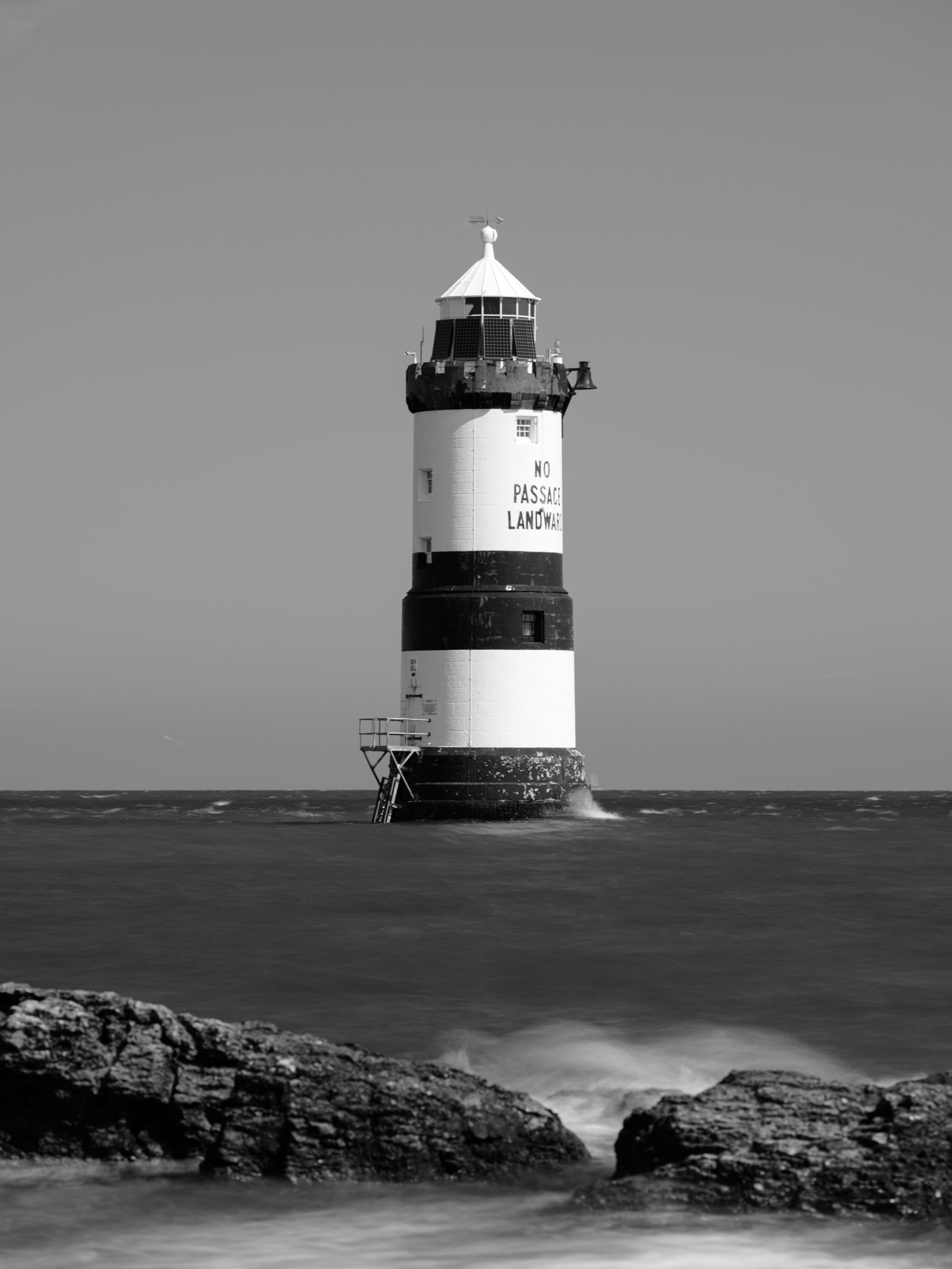 Black and white photo of a lighthouse standing in the sea, with rocks in the foreground and the sky in the background.
