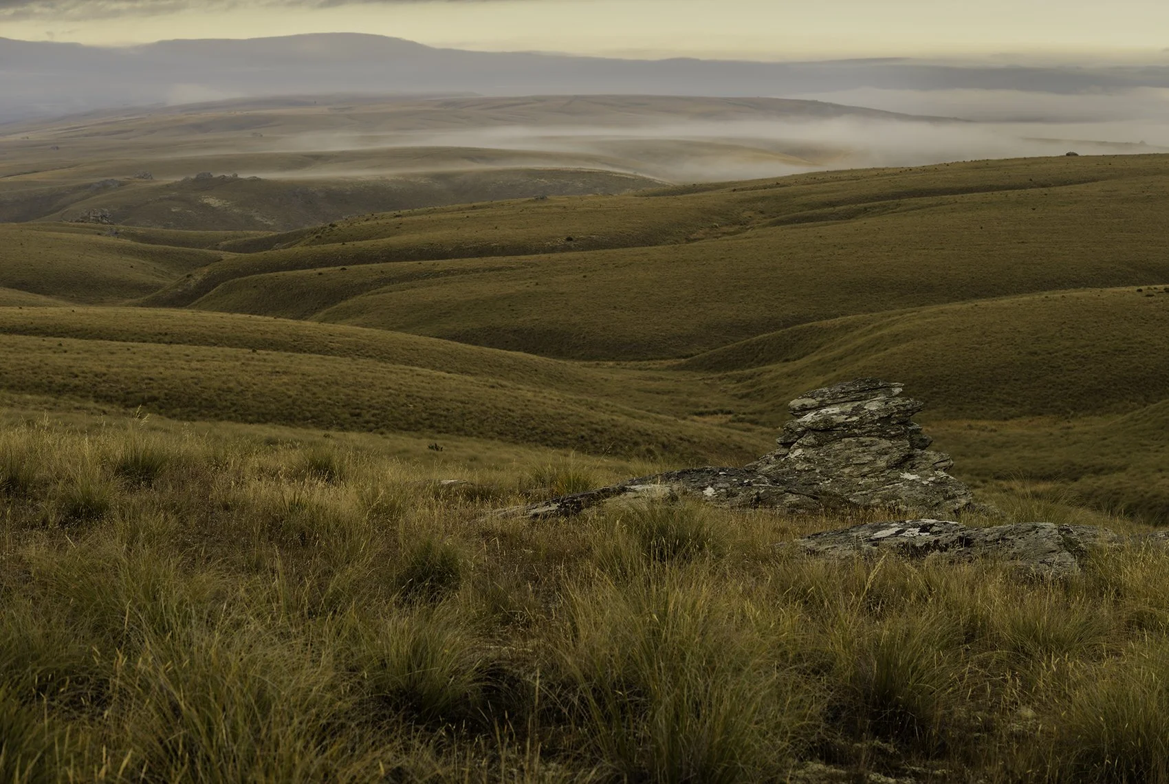 Rolling grass-covered hills with a stone formation in the foreground and misty clouds in the distance.