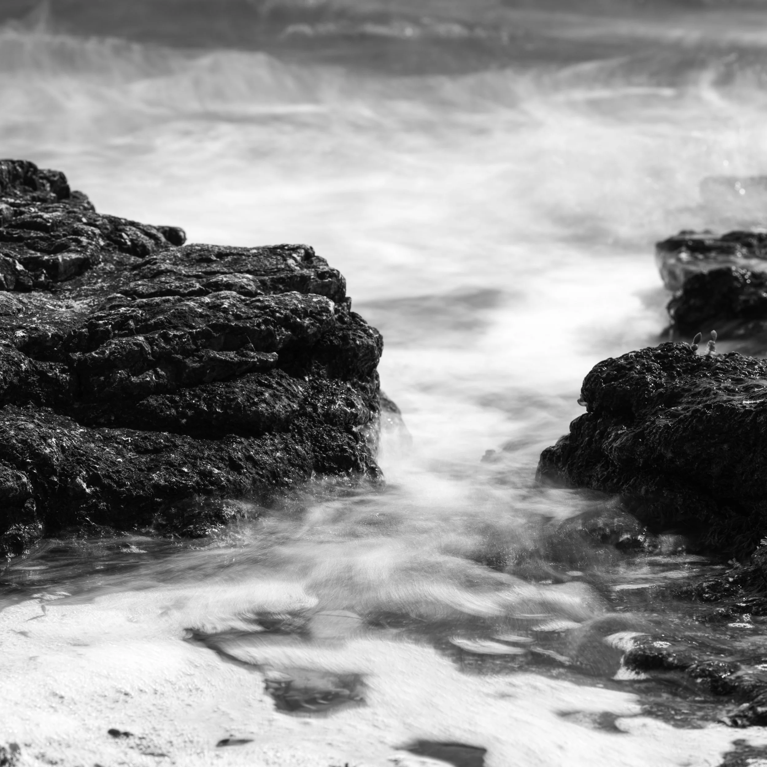 Black and white photo of ocean waves hitting rocky shoreline with sandy beach in foreground.