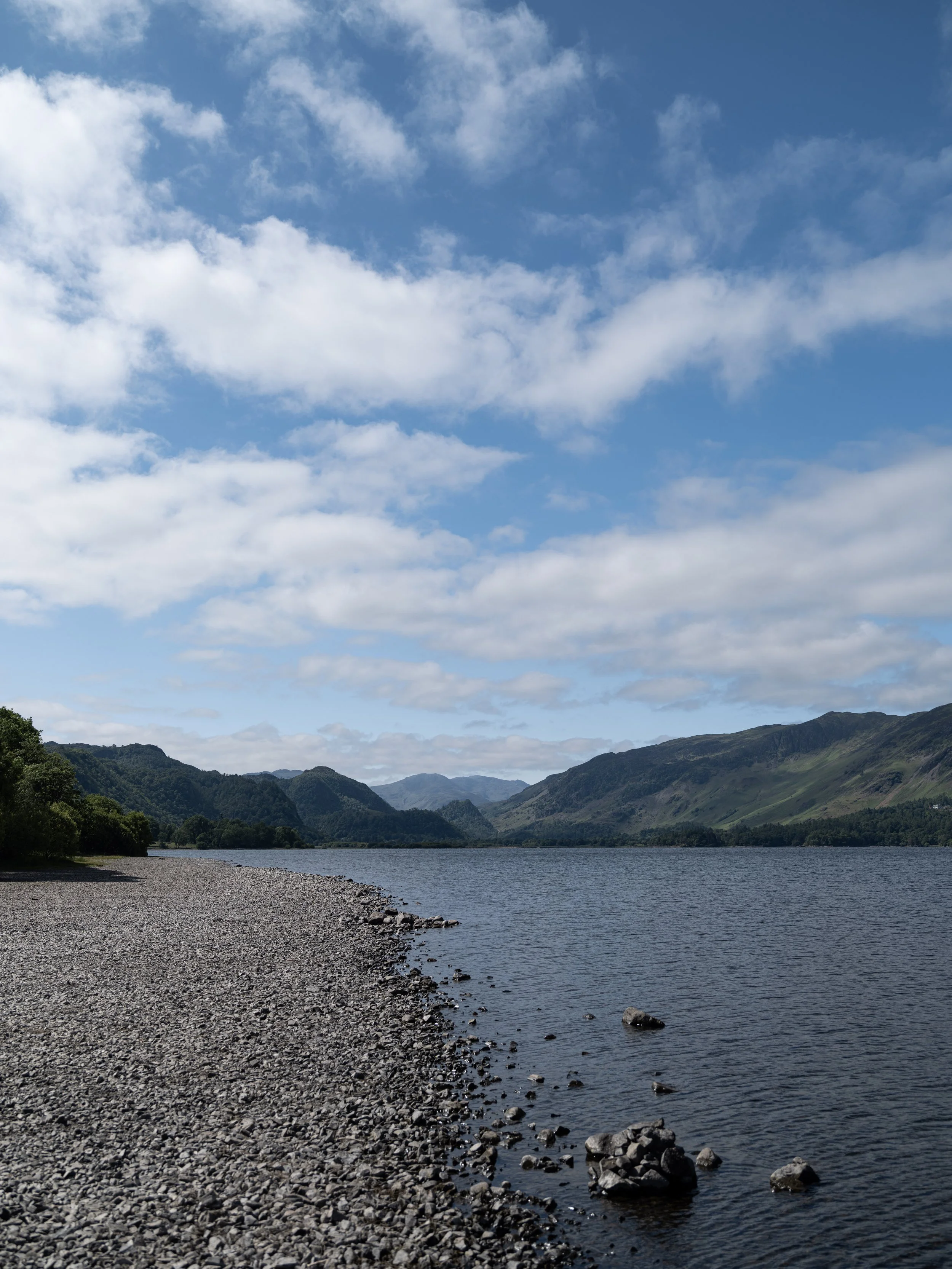 Scenic view of a lake with a rocky shoreline, surrounded by green mountains under a partly cloudy blue sky.