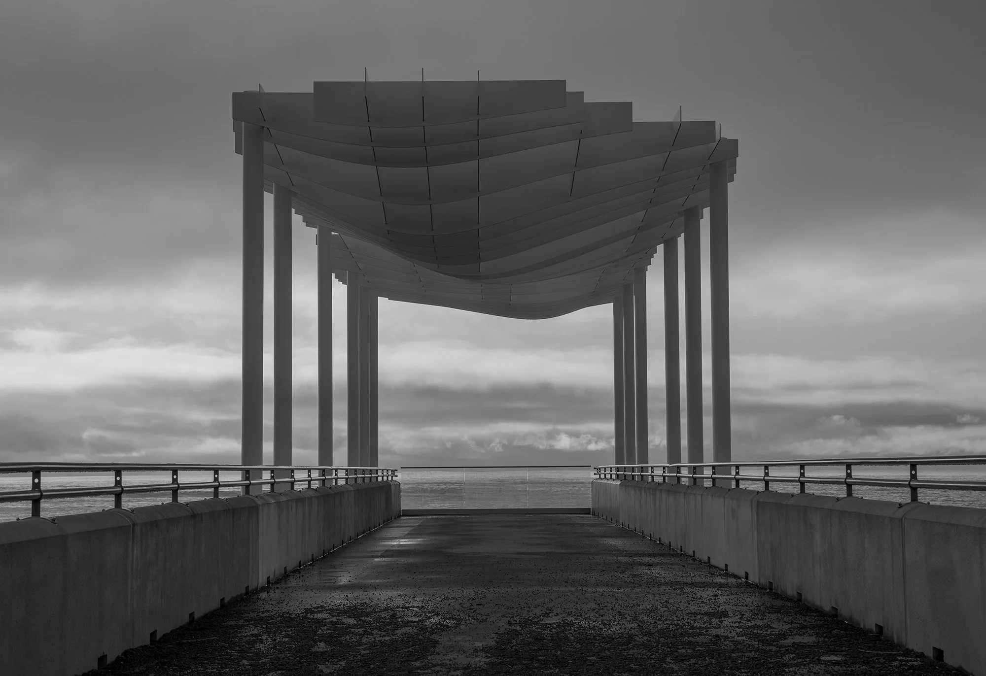 Black and white photo of a modern pier or walkway leading to a large, uniquely shaped, covered structure over the water with an overcast sky.