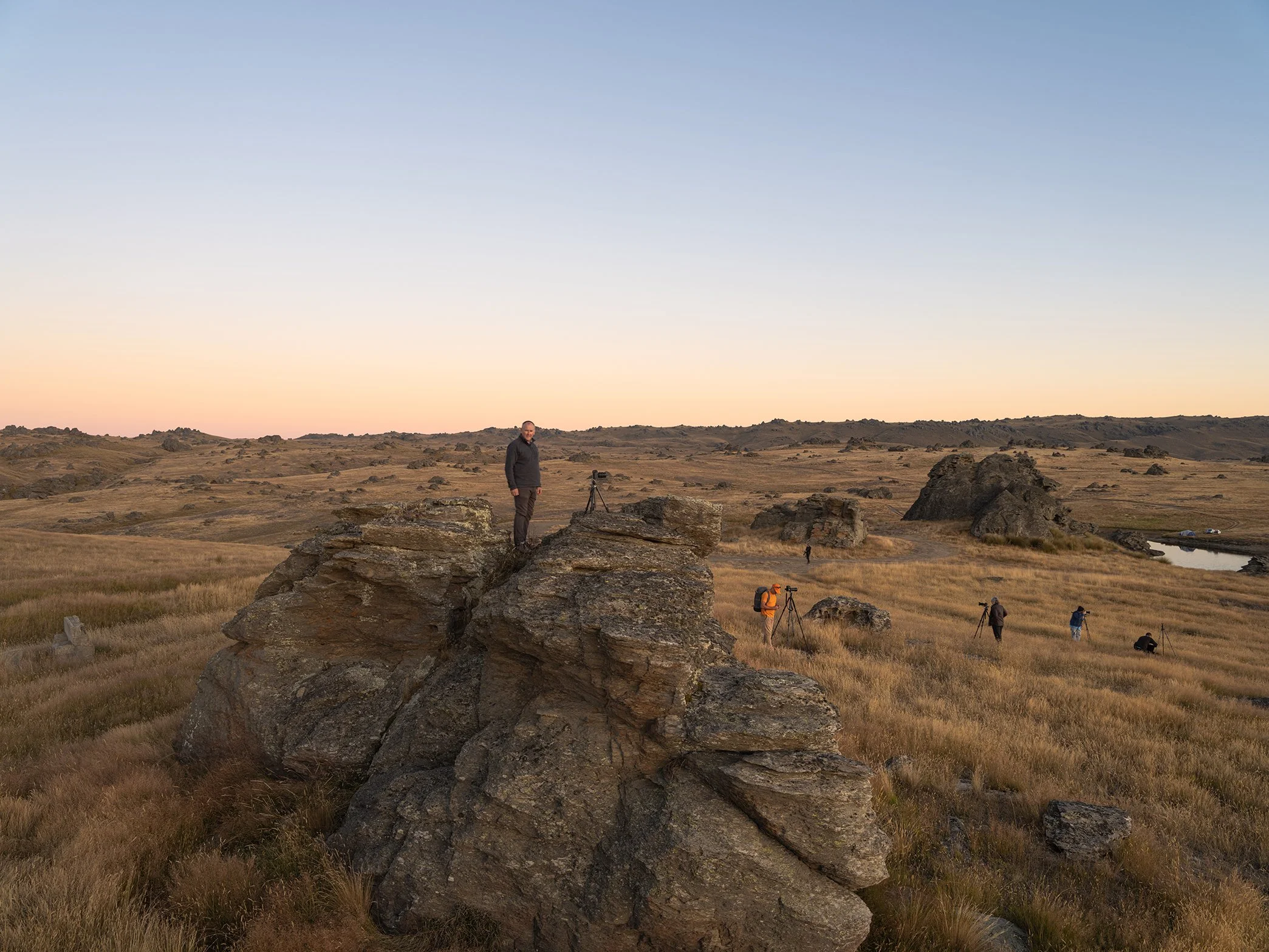 A group of photographers capturing the landscape at sunset in a grassy, rocky field with hills in the background.