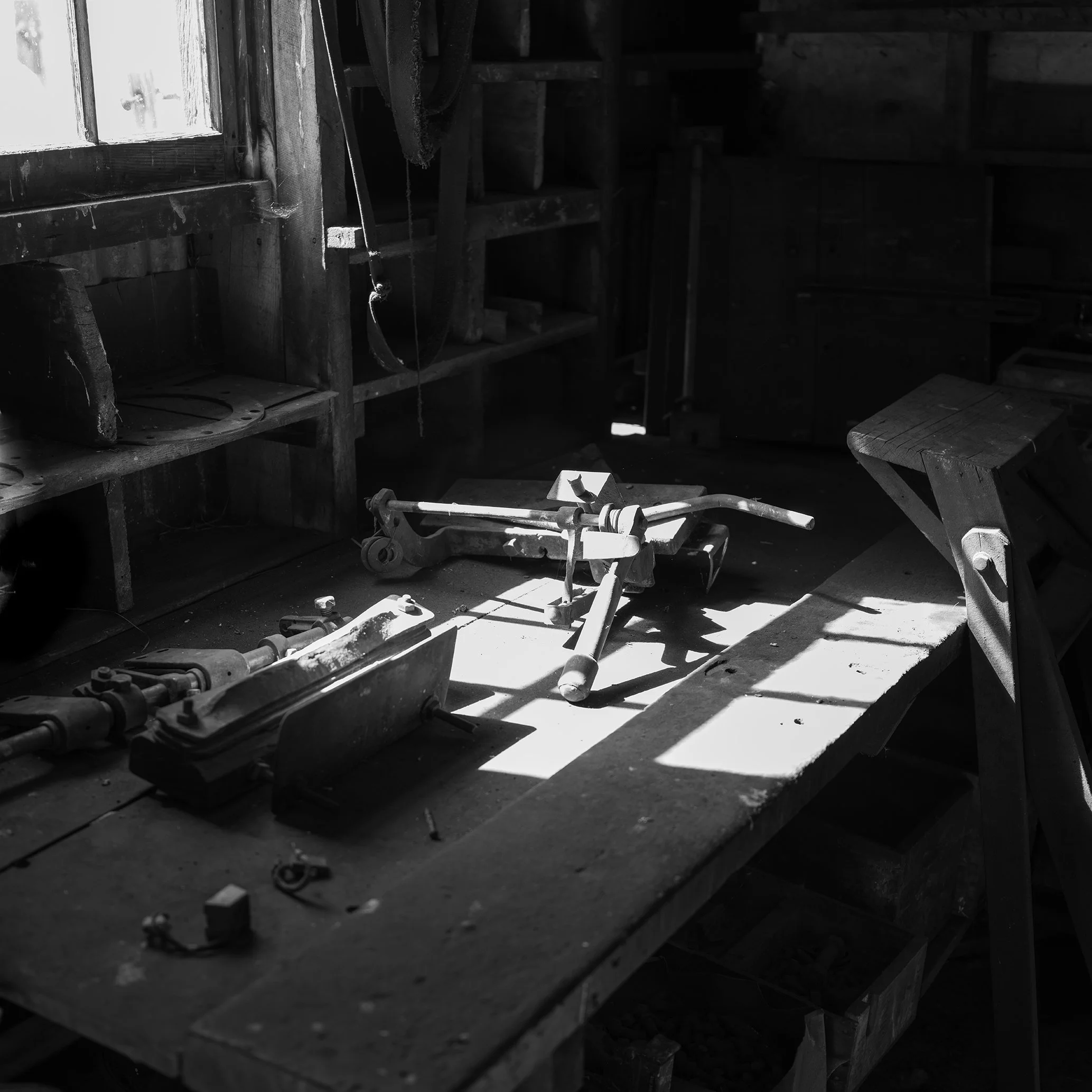 Old woodworking tools and equipment on a dusty workbench in a dimly lit workshop with sunlight streaming through a window.