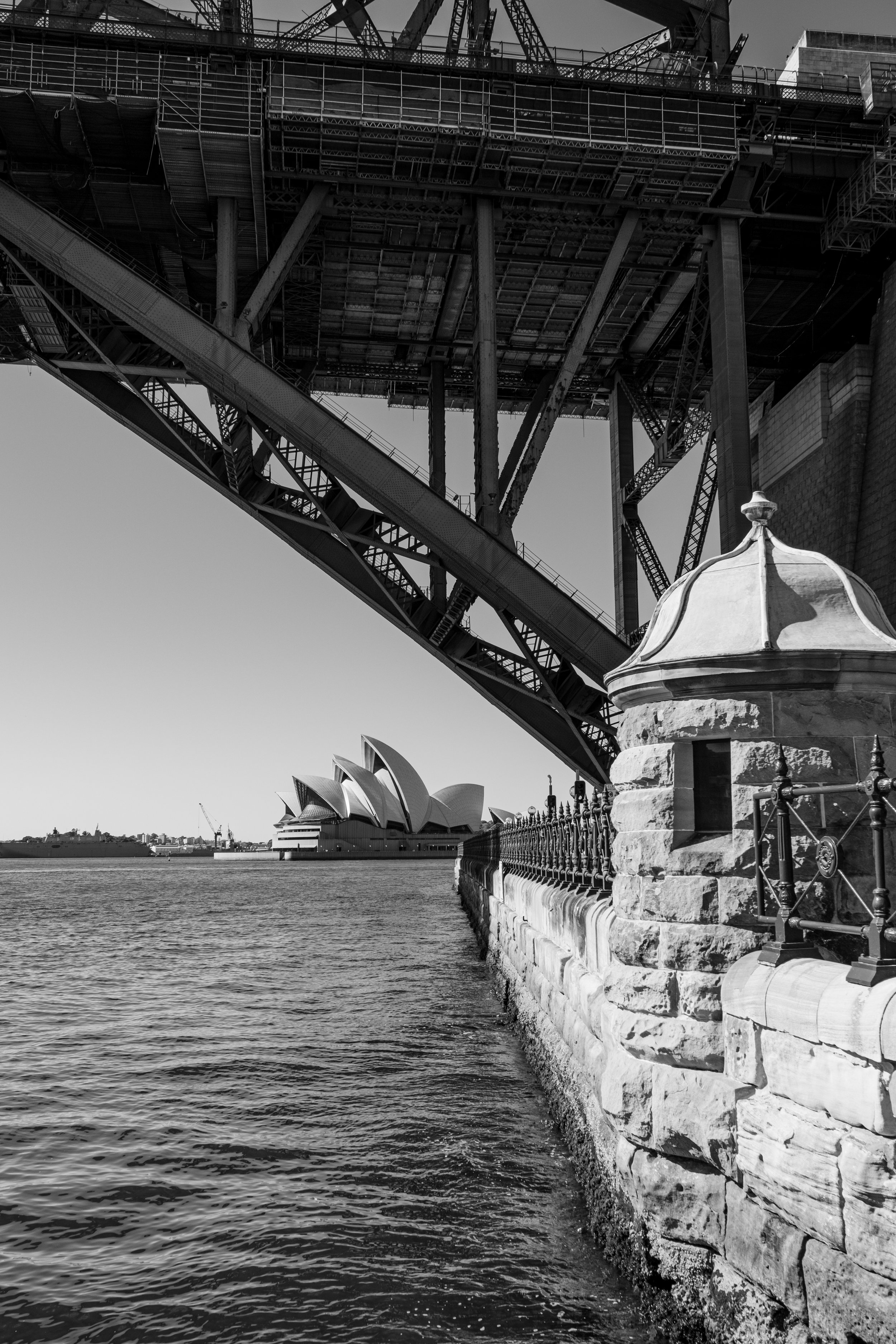 Black and white photo of the Sydney Harbour Bridge in the foreground, with the Sydney Opera House visible in the distance across the water.