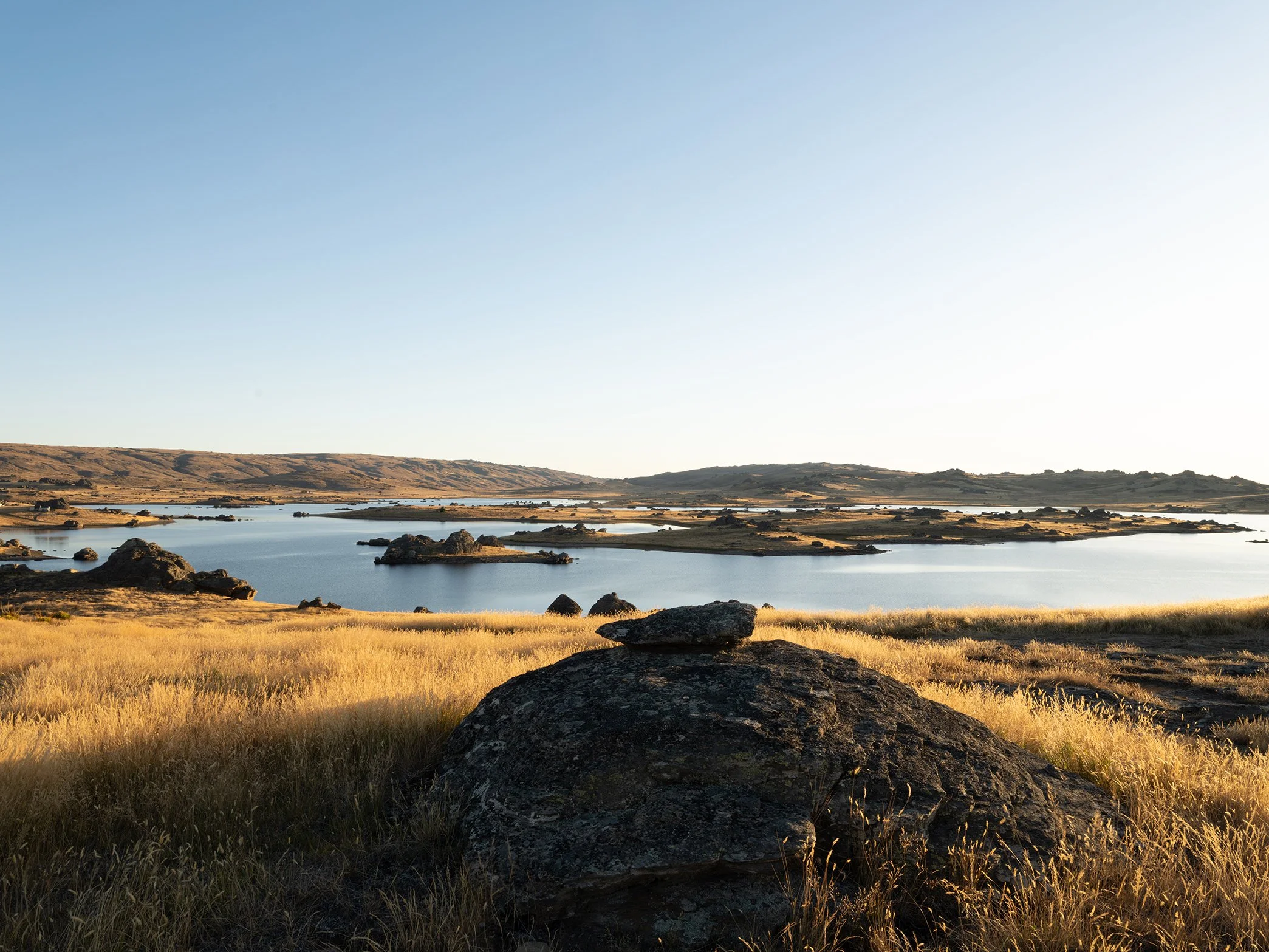 Serene landscape of a lake with small islands and rocky formations, surrounded by grassy fields under a clear sky