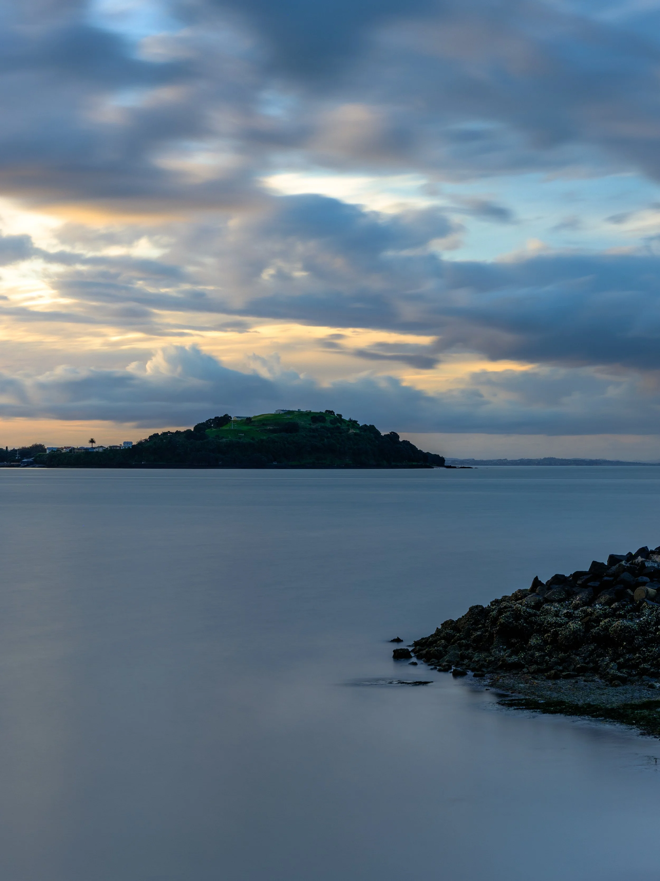 A calm body of water with an island in the distance under a cloudy sky at sunset.