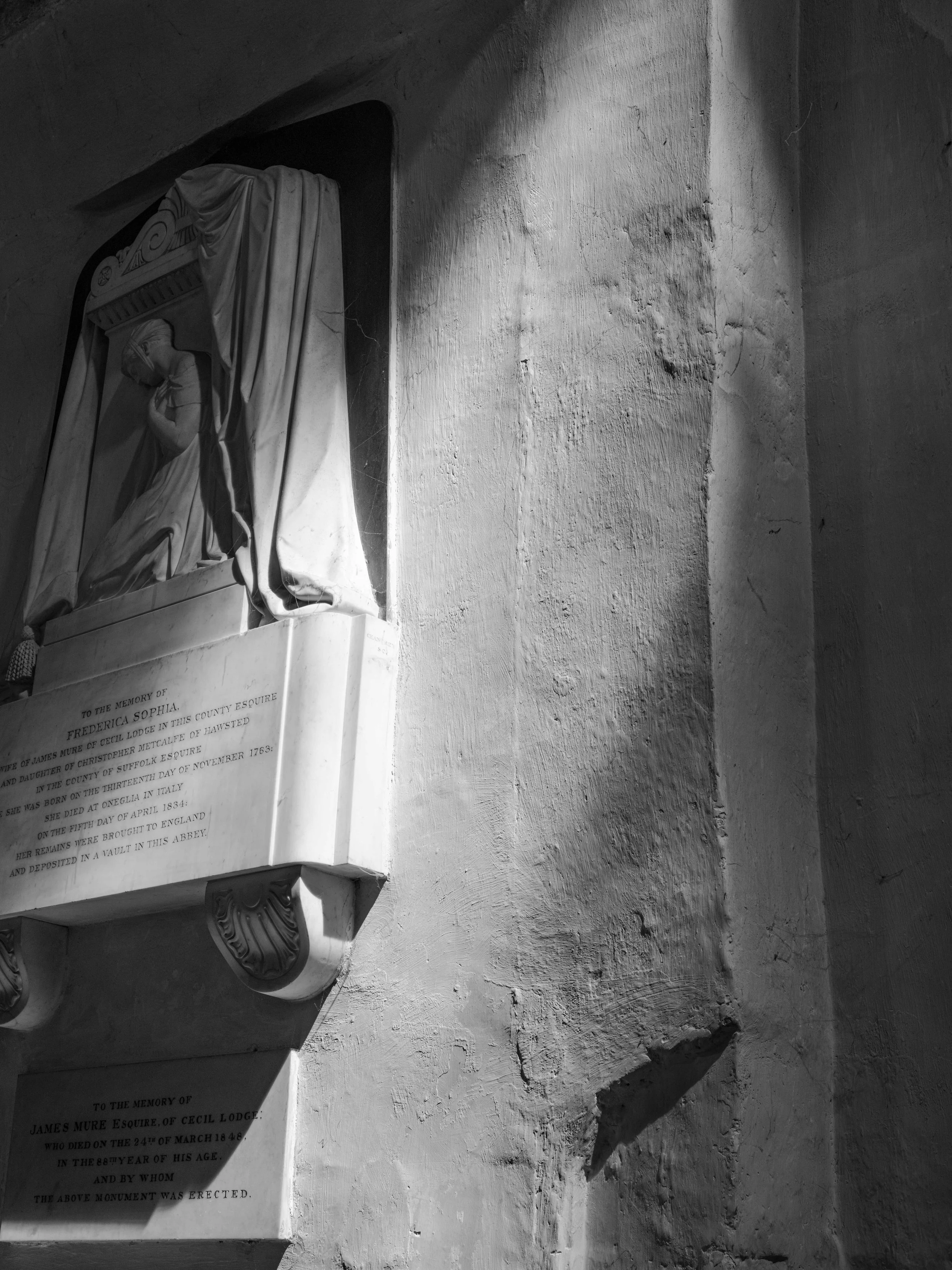 Black and white photograph of a religious relief sculpture on a wall, featuring a figure kneeling and praying under a canopy. The sculpture is set into a small alcove and is accompanied by a plaque with engraved text.