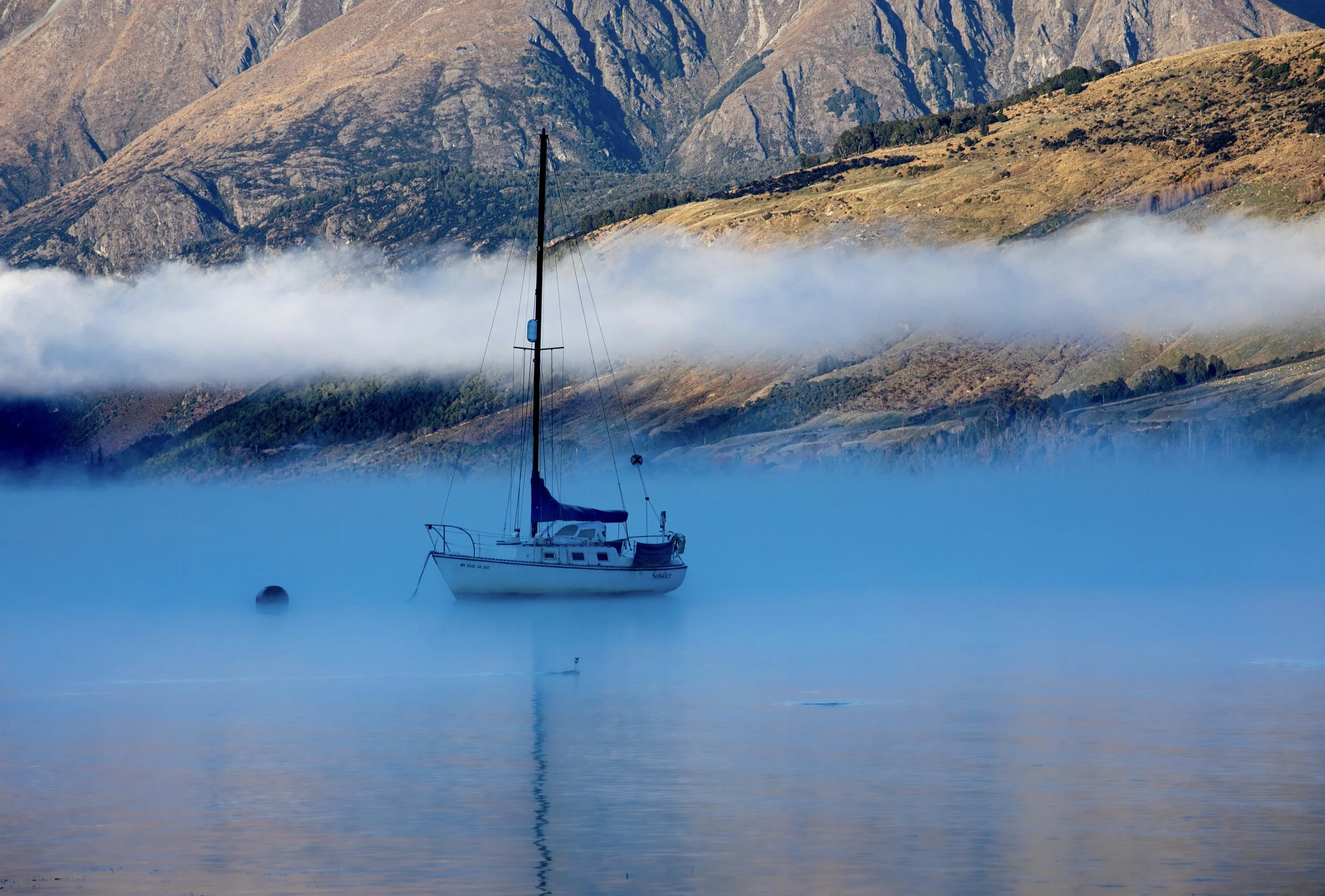A sailboat anchored in calm, foggy water with mountains in the background and a line of mist crossing the hills.