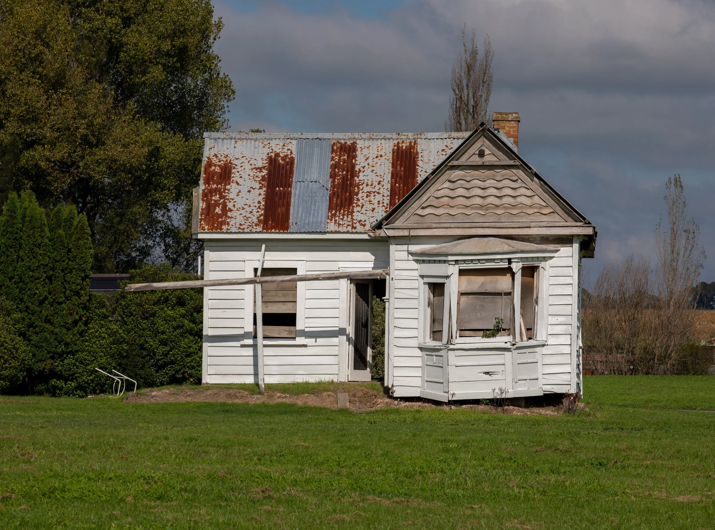 Old, abandoned house with a rusty metal roof, boarded-up windows, and weathered wood siding surrounded by green grass and trees.