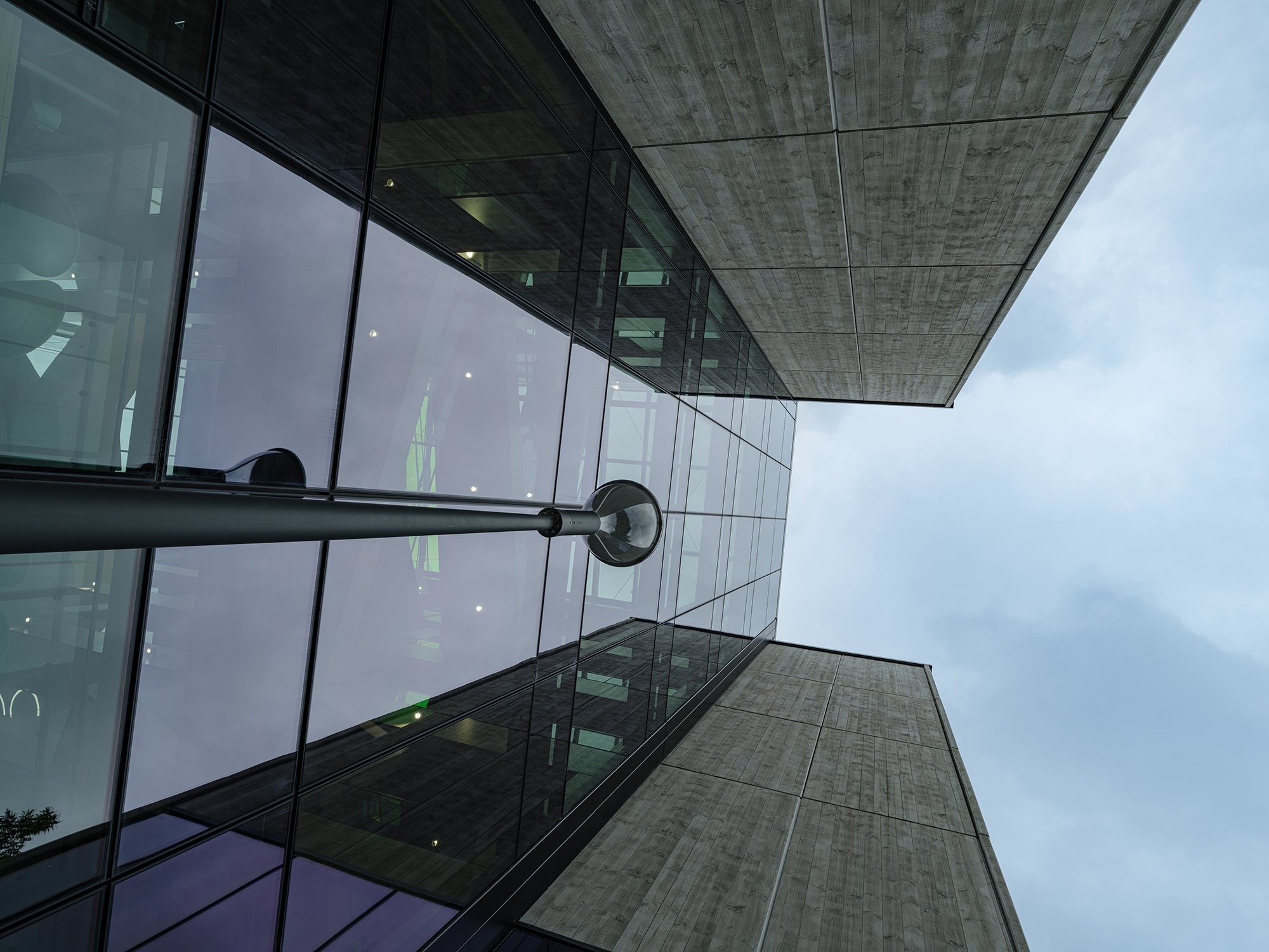 Looking up at a modern building with glass windows and concrete walls, featuring a street lamp and a cloudy sky.