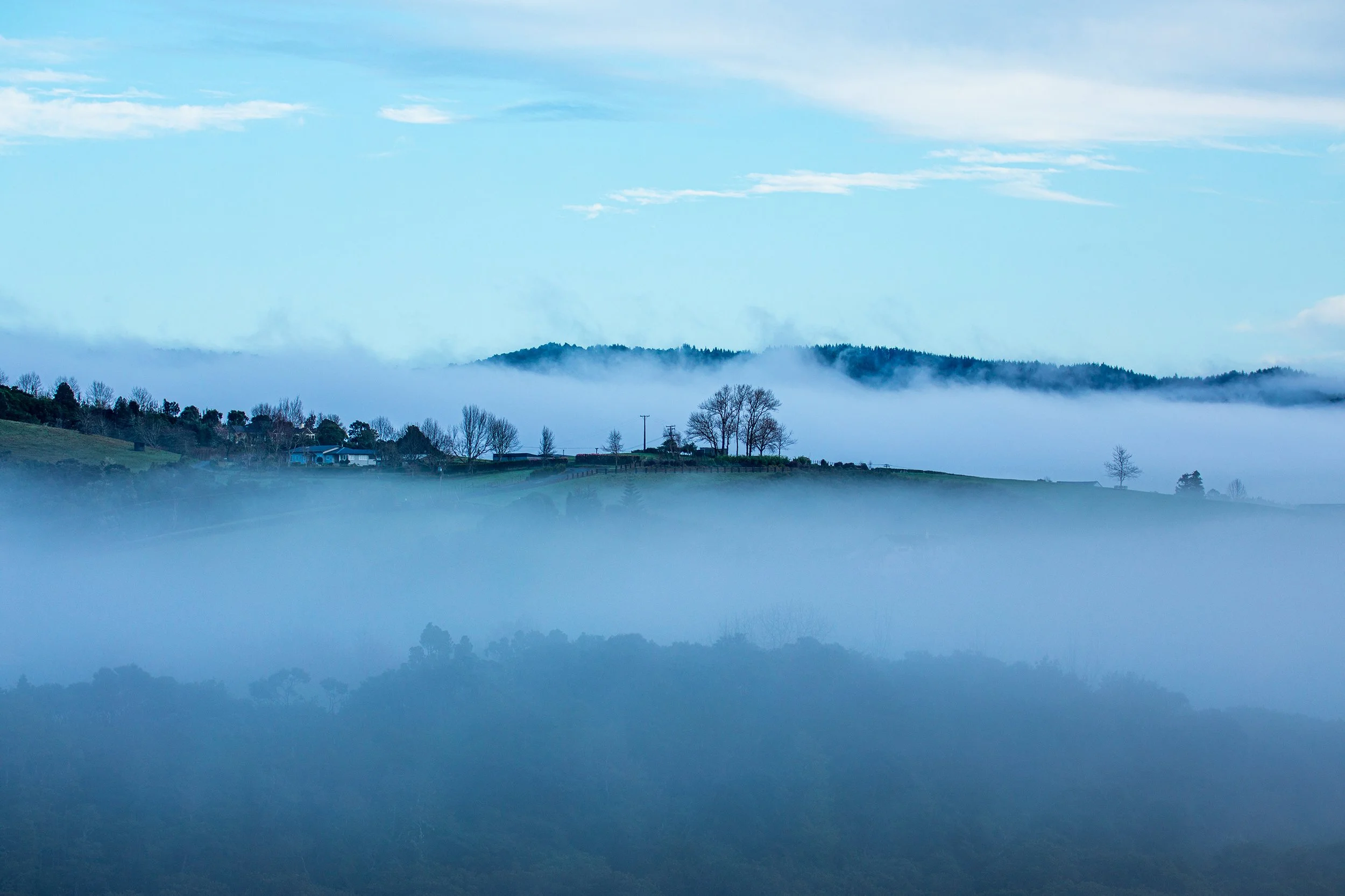 A landscape of rolling hills and trees partly covered in fog, with a blue sky and some clouds overhead.