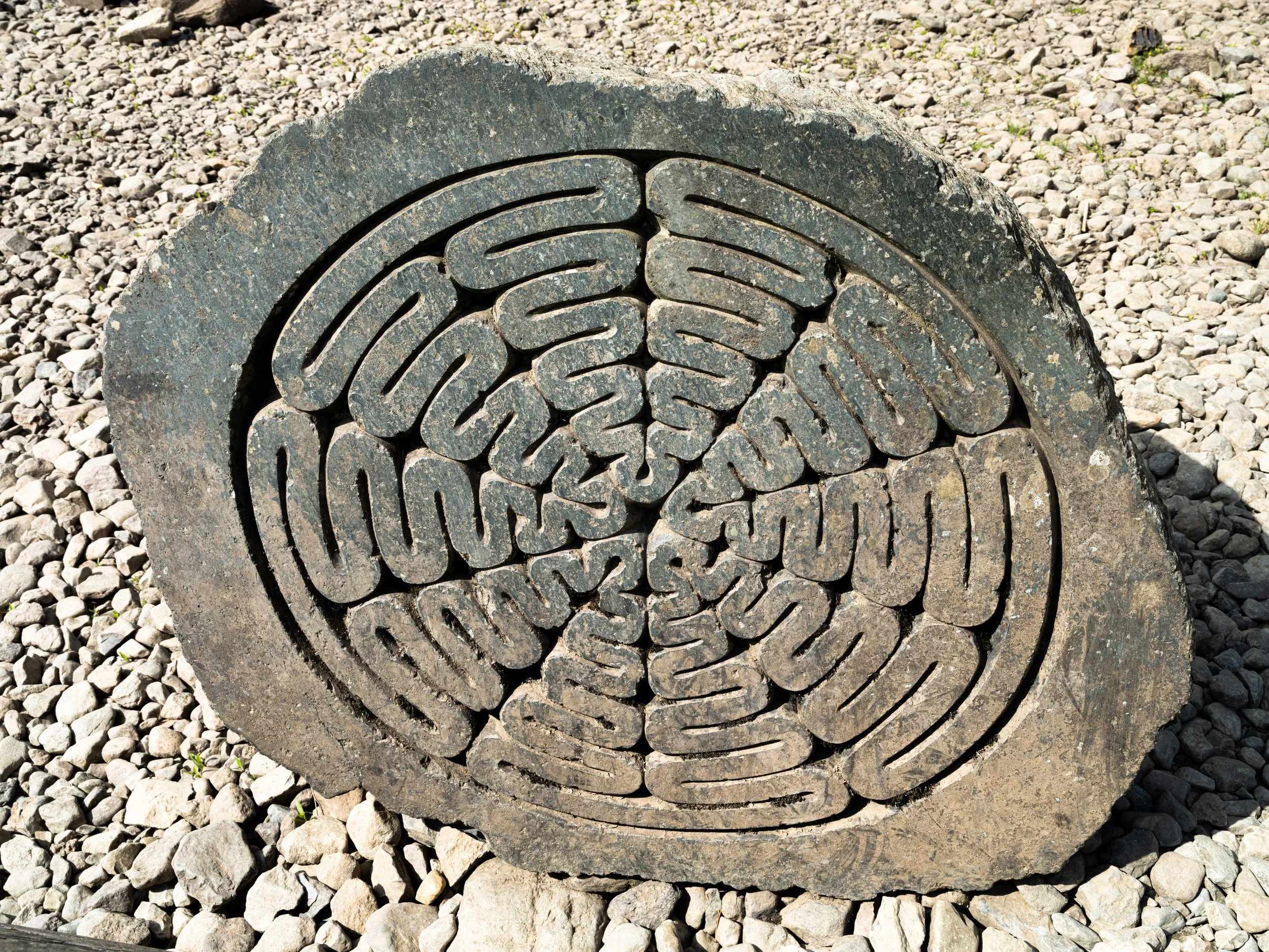 Round stone with carved intricate geometric pattern, placed on gravel surface.