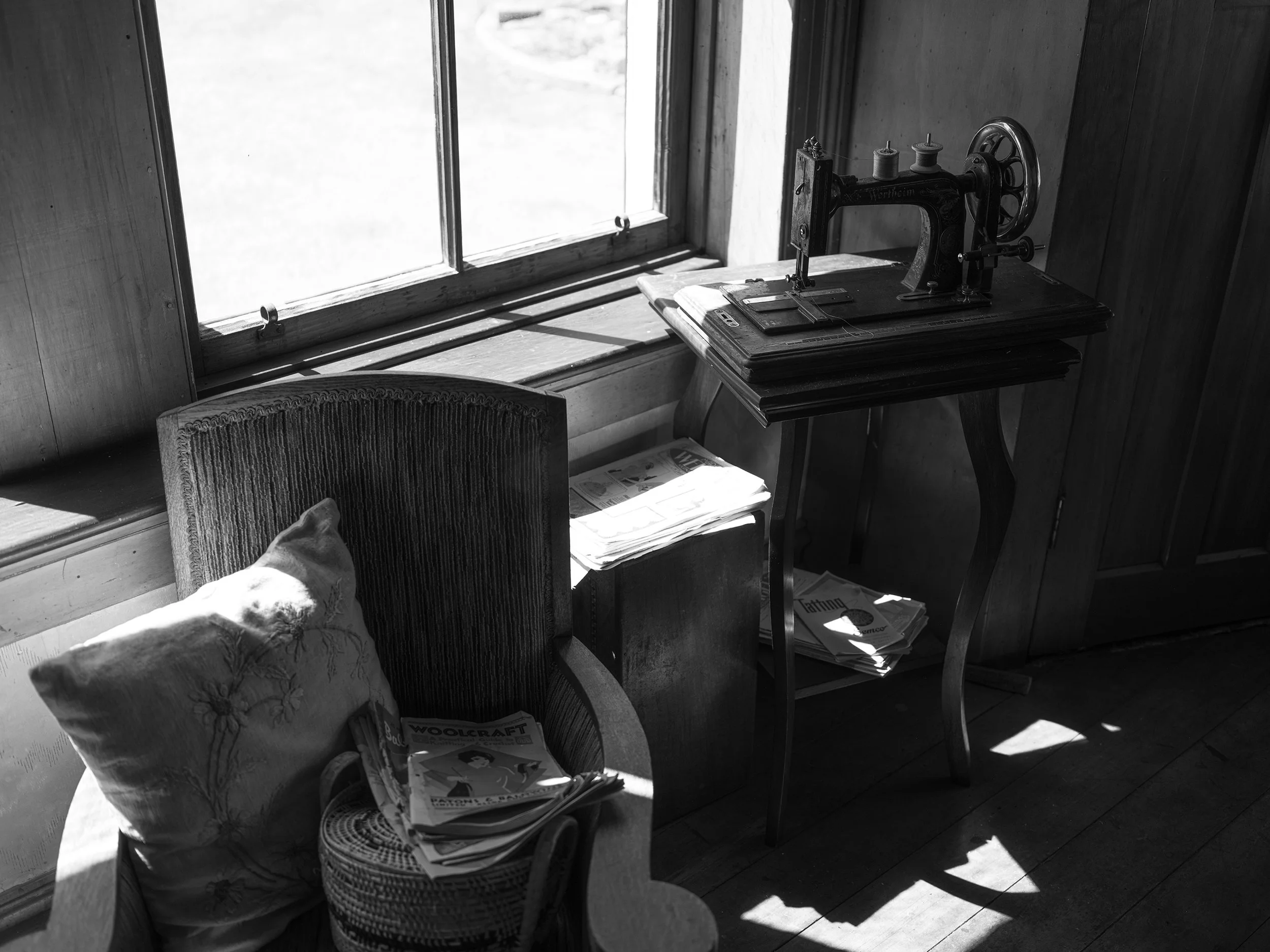 A vintage sewing machine on a wooden table next to a chair with a cushion, under a window with sunlight streaming in, storing newspapers underneath.
