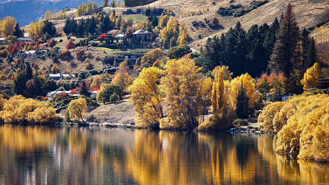 A scenic lakeside landscape with trees displaying fall colors, reflecting in calm water, and houses on the hillside in the background.