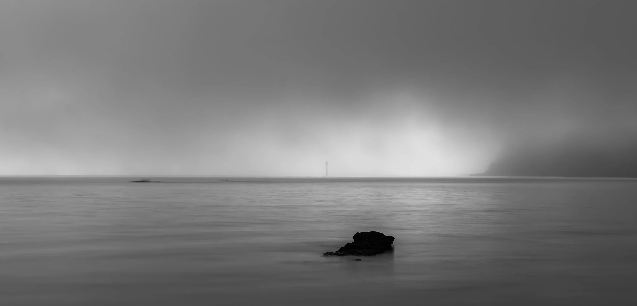 A black and white photo of a calm ocean with a small rock in the foreground, a distant lighthouse or tower on the horizon, and a cloudy sky.