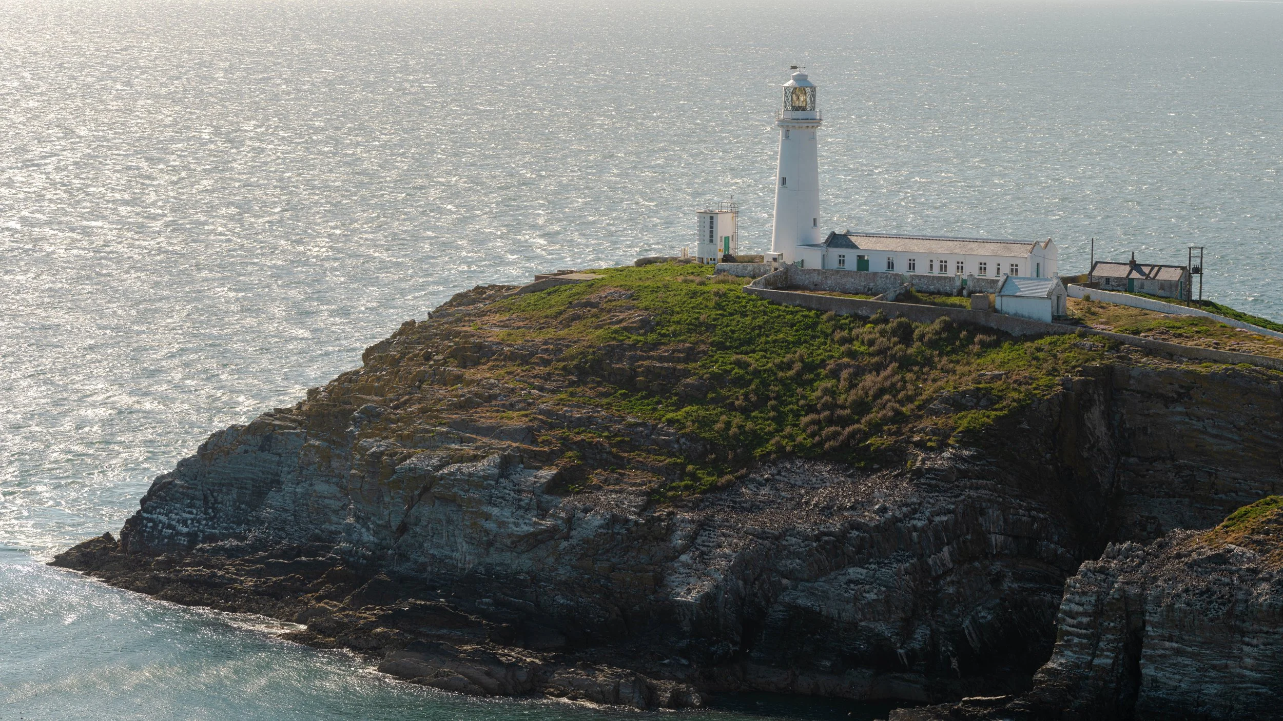 A white lighthouse on a grassy cliff overlooking the ocean with a partly cloudy sky.