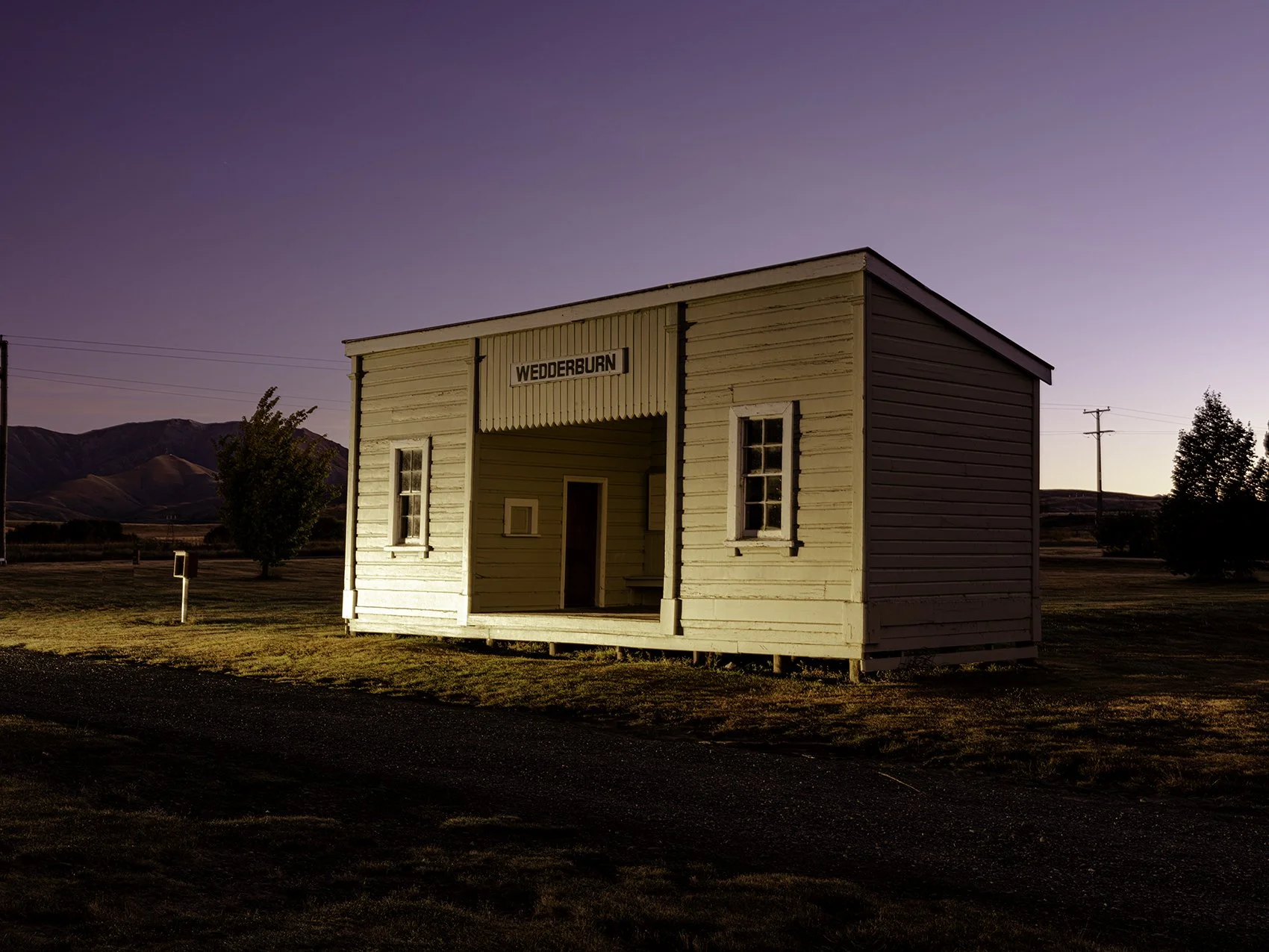 Old wooden train station building labeled 'Wedderburn' in a rural landscape with mountains in the background at sunset