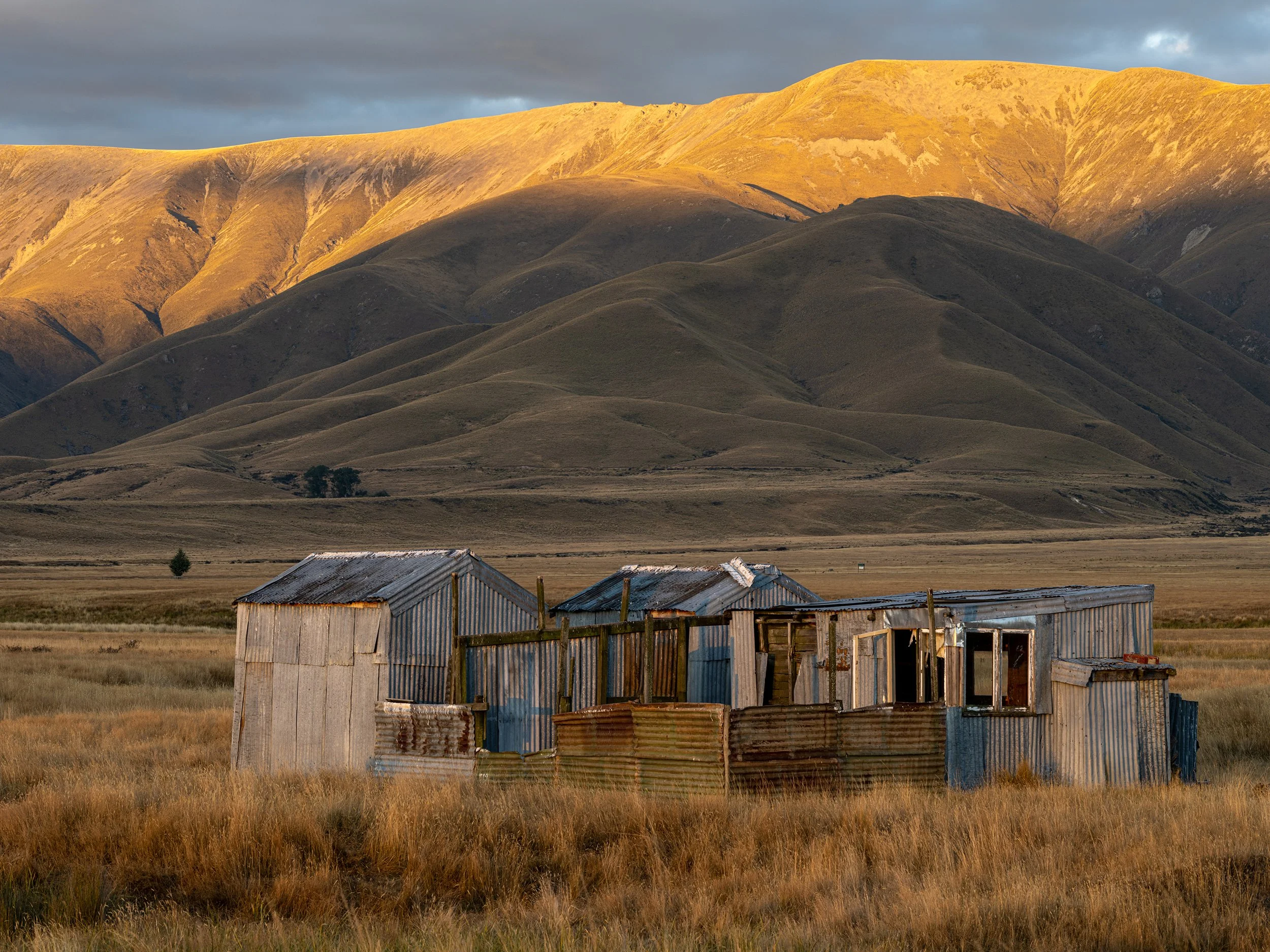 Abandoned metal and wooden shed in a grassy field with rolling hills and mountains in the background under a cloudy sky.