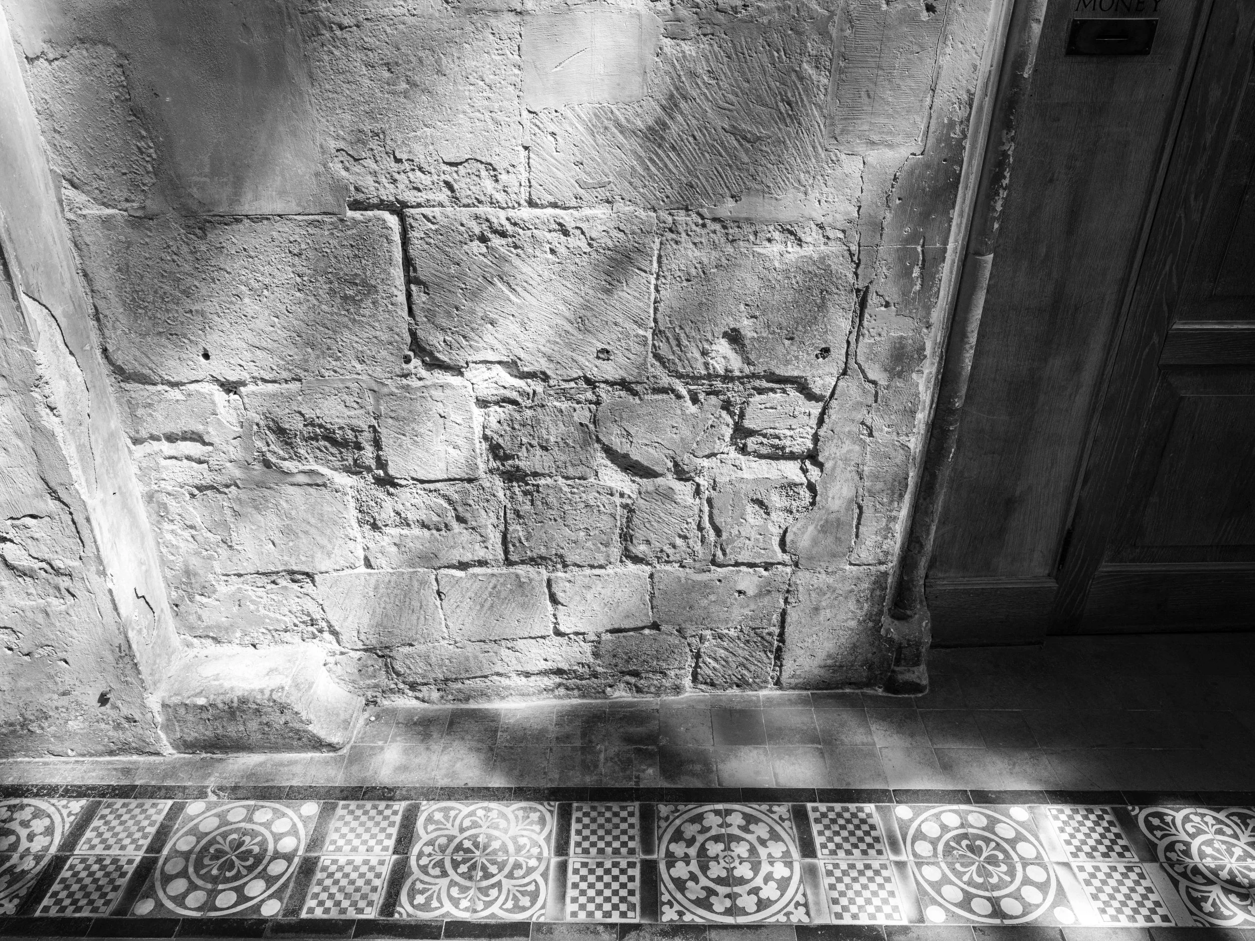 A black and white photo showing a weathered, exposed brick wall adjacent to a wooden cabinet and patterned tile flooring.