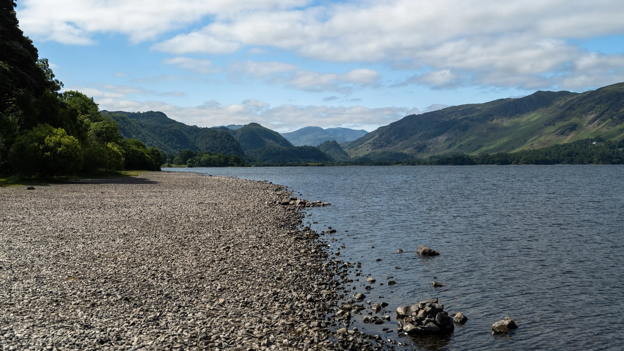 A lakeside pebbled beach with green trees, mountains in the distance, partly cloudy sky.