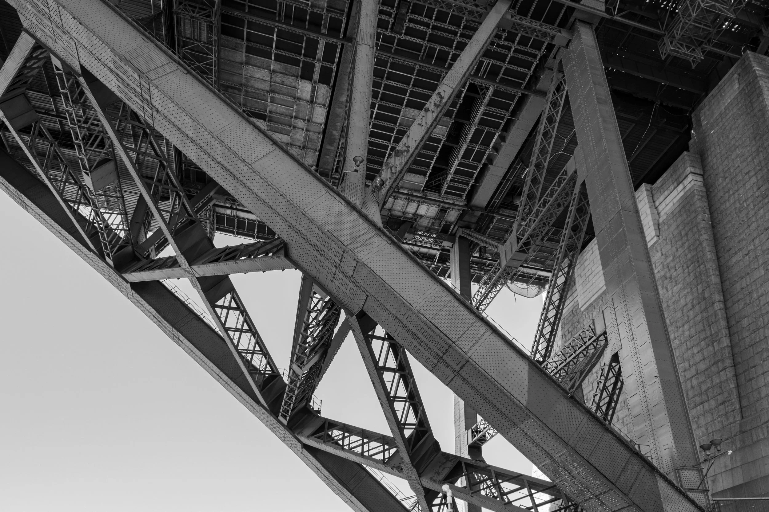 Black and white photograph of the underside of a large bridge showing steel beams, girders, and supporting structures.
