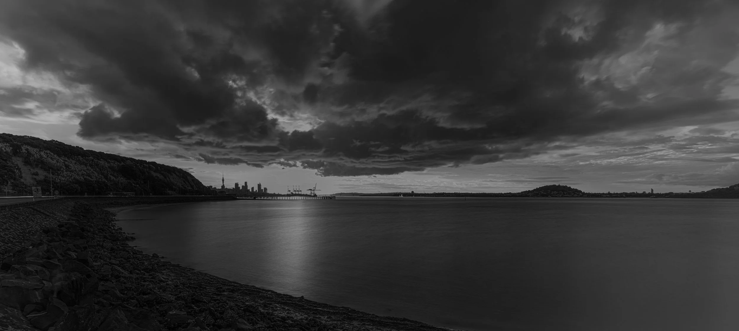 A black and white photo of a harbor with dark, cloudy skies and a city skyline in the distance.