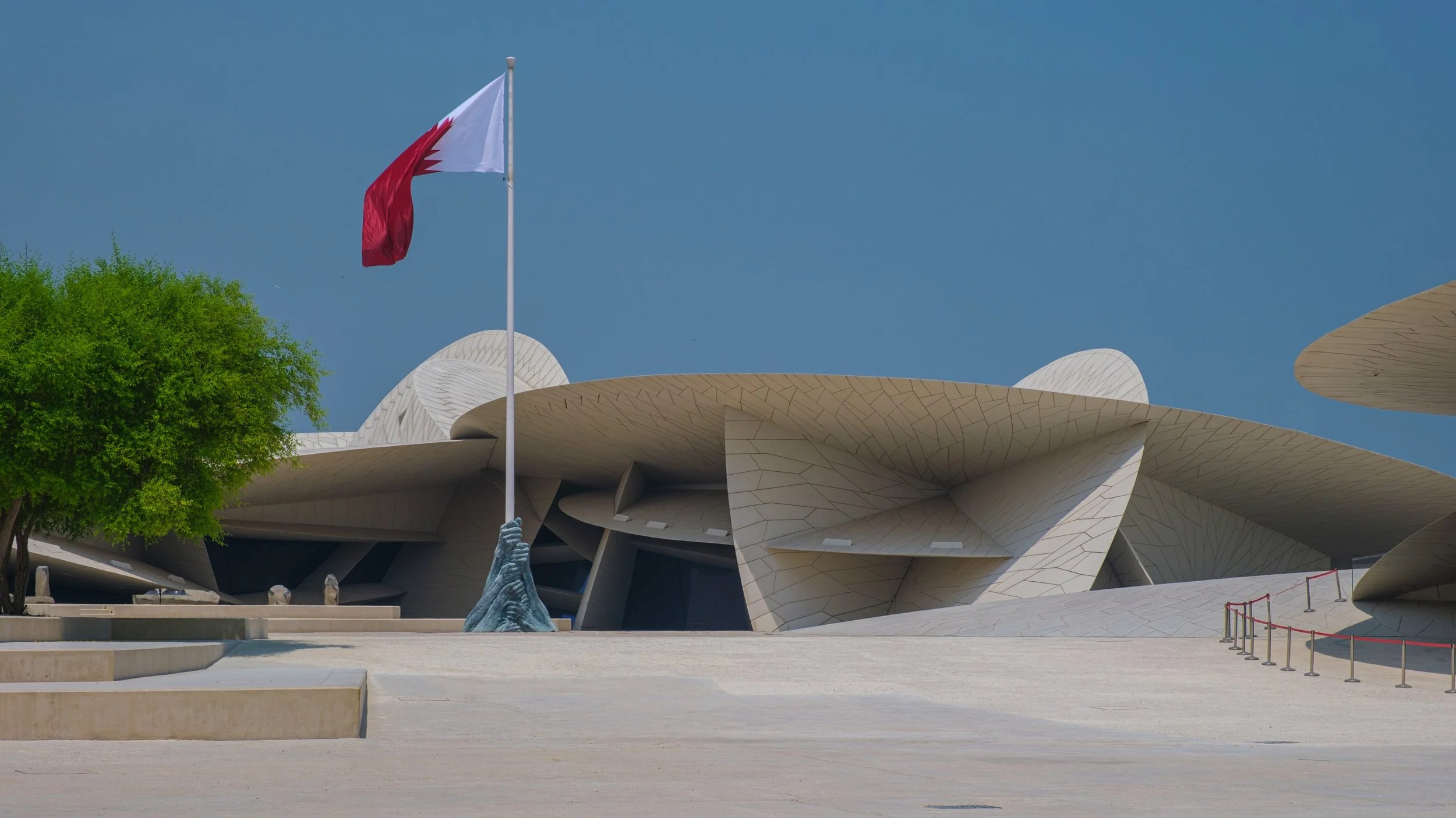 Modern architectural building with large, curved, white panels under a blue sky, a green tree on the left, and a flagpole with a red and white flag.