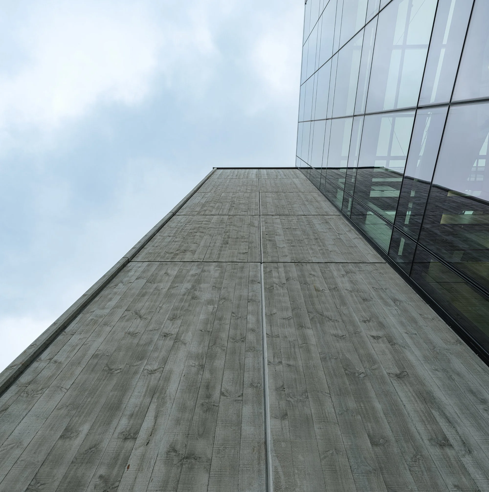 Looking up at the corner of a modern building with concrete and glass facade against a cloudy sky.